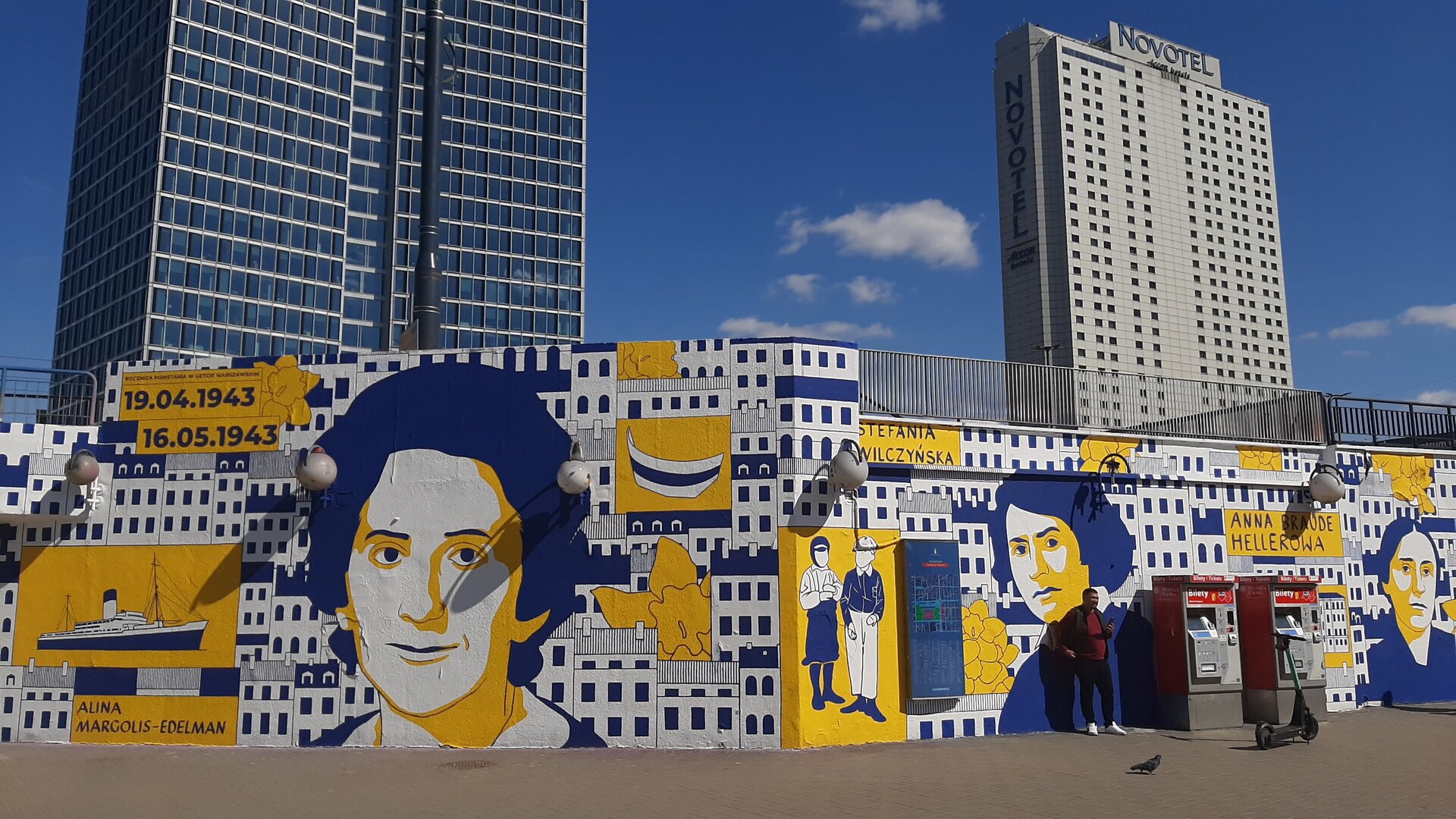 Zdjęcie przedstawia mural w kolorze żółtym i granatowym na białym tle. Najbardziej wyróżniające się elementy to twarze trzech kobiet, podpisane następującymi nazwiskami: Alina Margolis‑Edelman, Stefania Wilczyńska, Anna Braude Hellerowa. W tle namalowano bloki. Inne elementy to daty: 19.04.1943 oraz 16.05.1943, statek i żonkile. Mural znajduje się na murze, za którym stoją dwa wieżowce.