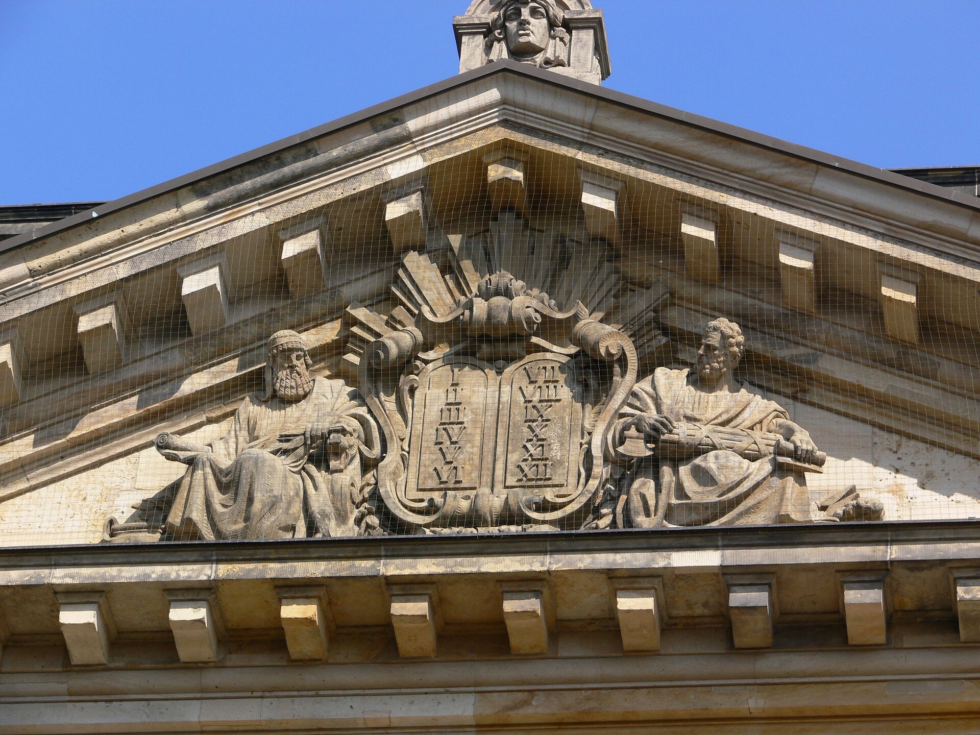 Ilustracja przedstawia fotografię „Detal z budynku Sądu Najwyższego w Lipsku”, której autorem jest Andreas Praefcke. Znajdują się na niej posągi dwóch siedzących mężczyzn o długich, kręconych włosach i brodzie. Ubrani są w długie szaty. Pomiędzy nimi są 2 zaokrąglone tablice. Na lewej tablicy są liczby rzymskie od I do VI, ułożone jedna pod drugą. Na prawej tablicy są liczby rzymskie od VII do XII, ułożone jedna pod drugą. Dookoła tablic są zdobienia. Detal z budynku znajduje się tuż pod dachem. Na dachu jest posąg przedstawiający twarz. W tle jest błękitne niebo.  