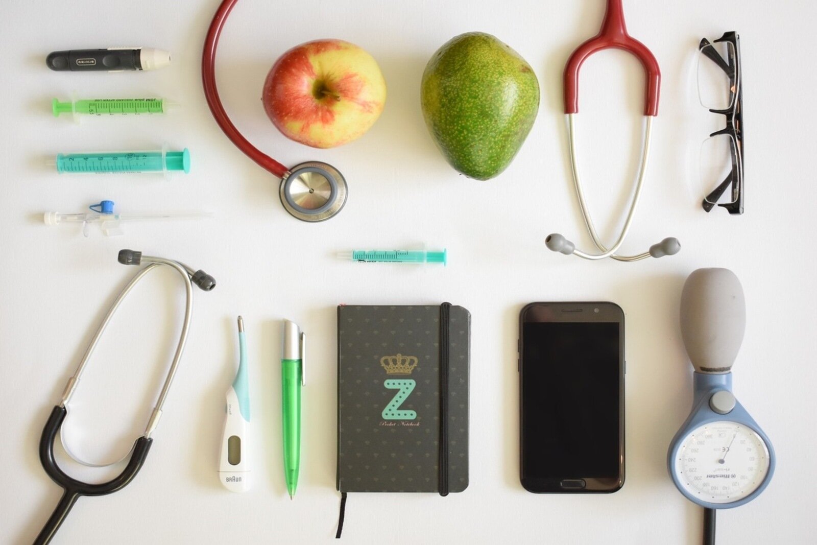 Zdjęcie przedstawia ułożone na stole przedmioty: strzykawki, stetoskop, termometr, okulary, miernik ciśnienia krwi, notes oraz jabłko i gruszka. 