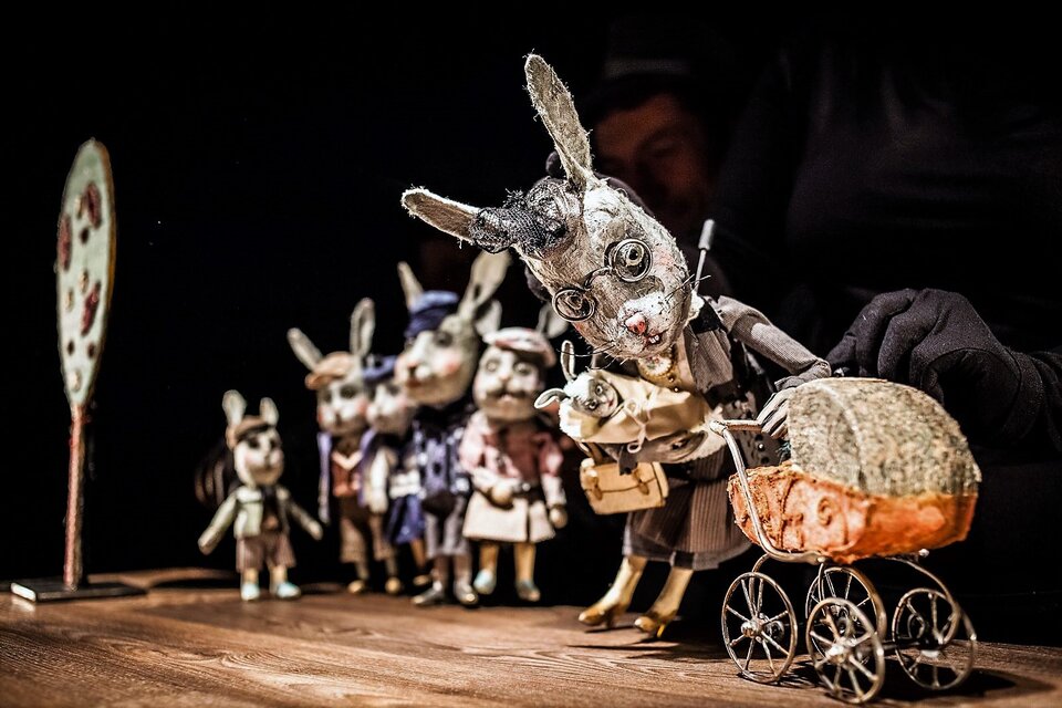 Ilustracja przedstawia scenę ze spektaklu "A niech to gęś kopnie", rozgrywającą się w teatrze lalkowym. Lalkami na scenie porusza aktor, którego postać jest prawie niewidoczna – widać jedynie jego rękę w czarnej rękawiczce, która animuje lalki. Na scenie znajduje się rodzina królików ustawiona jakby w kolejce. Na przodzie stoi mama‑królik, trzymająca dziecko w beciku, pochylona nad drugim dzieckiem siedzącym w wózku. Za nią stoją zarówno małe, jak i duże króliki, wszystkie elegancko ubrane, przypominające ludzi, skierowane wzrokiem w stronę matki.