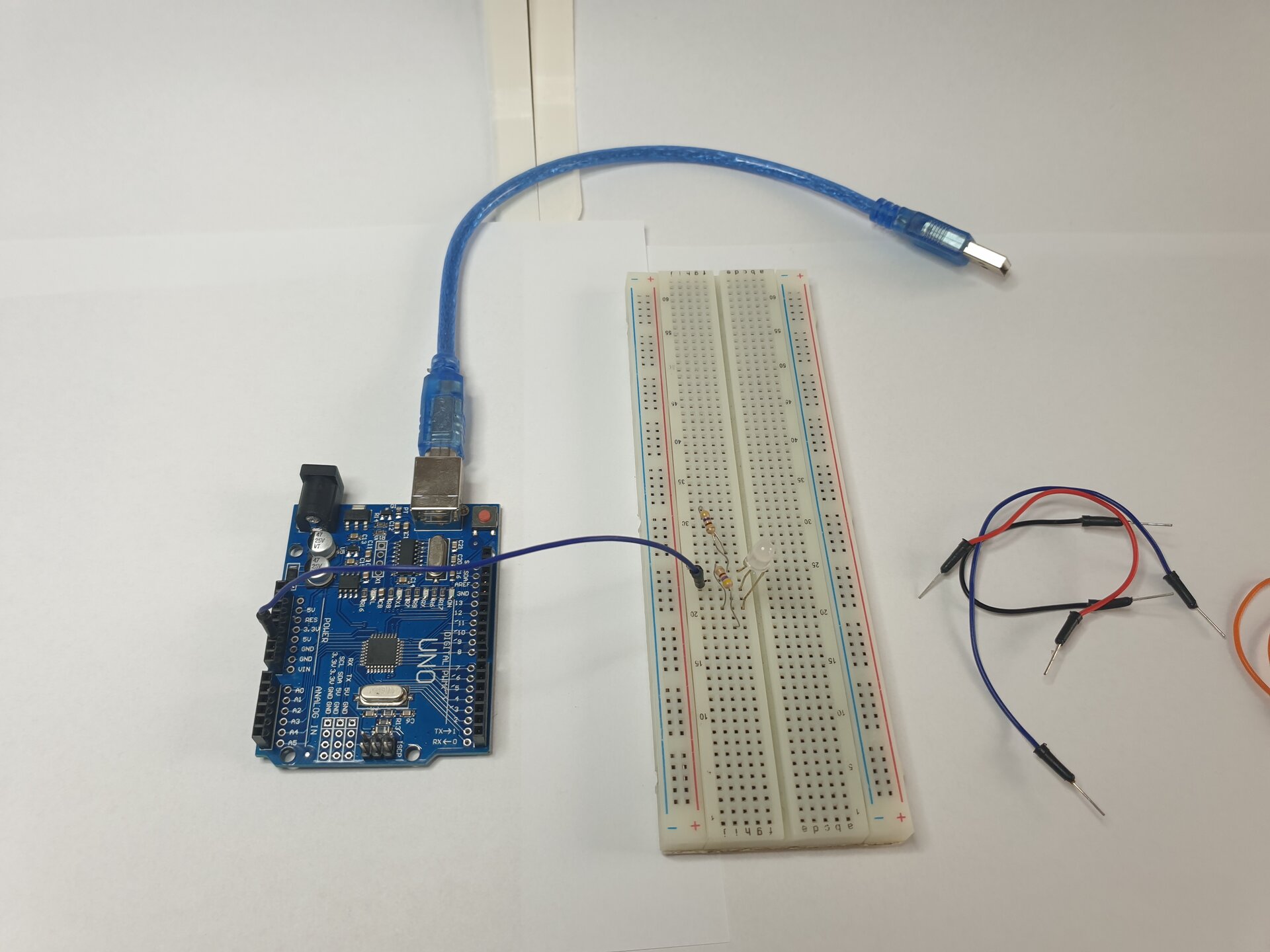Zdjęcie przedstawia moduł Arduino z podłączonym do niego kablem USB, płytkę stykową, w którą wpięto diodę oraz leżące obok płytki przewody. Połączono moduł z płytką za pomocą przewodu wpiętego w module w 5 V i w płytce w tym samym wierszu, co środkowa nóżka diody. Dwa rezystory wpięto w płytkę stykową w taki sposób, że jedna nóżka każdego rezystora jest w tym samym wierszu, co zewnętrzne nóżki diody, a druga nóżka każdego rezystora jest wpięta „na zewnątrz” nóżek diody.
