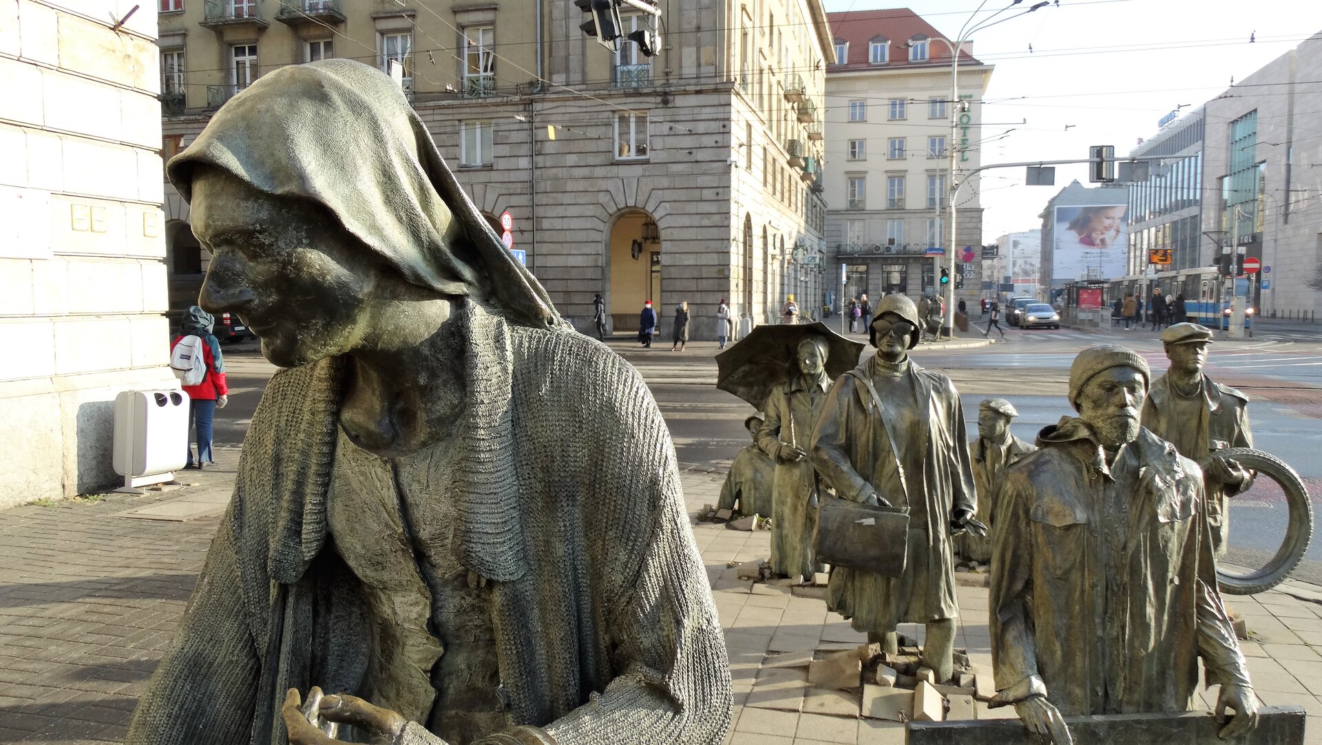 Zdjęcie wykonane w jasny słoneczny dzień przedstawia instalację - grupę naturalnej wielkości rzeźb postaci, które wychodzą z chodnika. Każda z rzeźb ustawiona jest w tę samą stronę. Każda z rzeźb przedstawia inną osobę. Najbliżej znajduje się kobieta w swetrze. Kolejny jest mężczyzna z brodą i czapką. Dalej starsza kobieta, mężczyzna trzymający oponę, kobieta z parasolem i inni. Rzeźby są bardzo szczegółowe – na twarzach przechodniów można zaobserwować zarost, zmarszczki, mimikę oraz detale ubioru. W tle ulica, budynki i przechodnie.