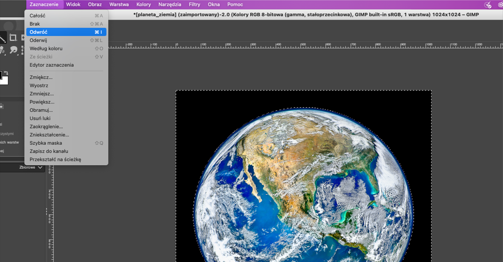 Ilustracja przedstawia wycinek okna w programie GIMP. Okno ma kolor szary, na górze i z lewej strony znajduje się podziałka z liczbami. Wewnątrz okna znajduje się fotografia – Ziemia widziana z kosmosu. Ma kształt okrągłej kuli z konturem Ameryki Północnej w kolorze brązowo – zielonym oraz białymi obłokami i niebieskim oceanem. Tło obrazka ma kolor czarny. Z szeregu ikonek z menu programu wybrana jest ikonka z symbolem różdżki. Ziemia obrysowana jest przerywaną, białą linią w kształcie koła, a cały obrazek białą, przerywaną linią tworzącą kształt kwadratu. W górnej części zdjęcia znajdują się zakładki menu programu Zaznaczenia, Widok, Obraz, Warstwa, Kolory, Narzędzia, Filtry, Okna oraz Pomoc. Wybrana została pozycja menu: Zaznaczenie, pod spodem widoczna jest rozwinięta, pionowa lista z pozycjami: Całość, Brak, Odwróć, Oderwij, Według koloru, Ze ścieżki, Edytor zaznaczenia, Zmiękcz.., Wyostrz, Zmniejsz…, Powiększ…, Obramuj…, Usuń łuki, Zaokrąglenie…, Zniekształcenie…, Szybka maska, Zapisz do kanału, Przekształć na ścieżkę. Z listy tej wybrana jest pozycja listy: Odwróć.  