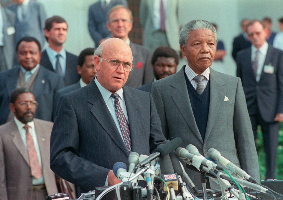 Zdjęcie przedstawia Nelsona Mandelę w towarzystwie Frederika de Klerk. Pierwszy z nich to czarnoskóry, starszy mężczyzna o jasnobrązowym odcieniu skóry. Ma on szeroki nos, niewielkie, ciemne oczy, na jego ustach widoczny jest uśmiech. Jego włosy są krótkie, szare oraz kręcone. Jest on ubrany w złotą, elegancką koszulę ze wzorzystym kołnierzykiem. Obok niego stoi Frederik de Klerk. Ma on wąski nos, cienkie wargi oraz ciemne oczy i złote, druciane okulary. Posiada łysiejącą głowę, a po bokach i z tyłu siwe włosy. Ubrany jest on w garnitur, koszulę i krawat pod szyją. Obaj stoją na ambonie, przed sobą mają mikrofony różnych stacji telewizyjnych i radiowych. W tle widać kilku mężczyzn białych i czarnych przysłuchujących się wystąpieniu.