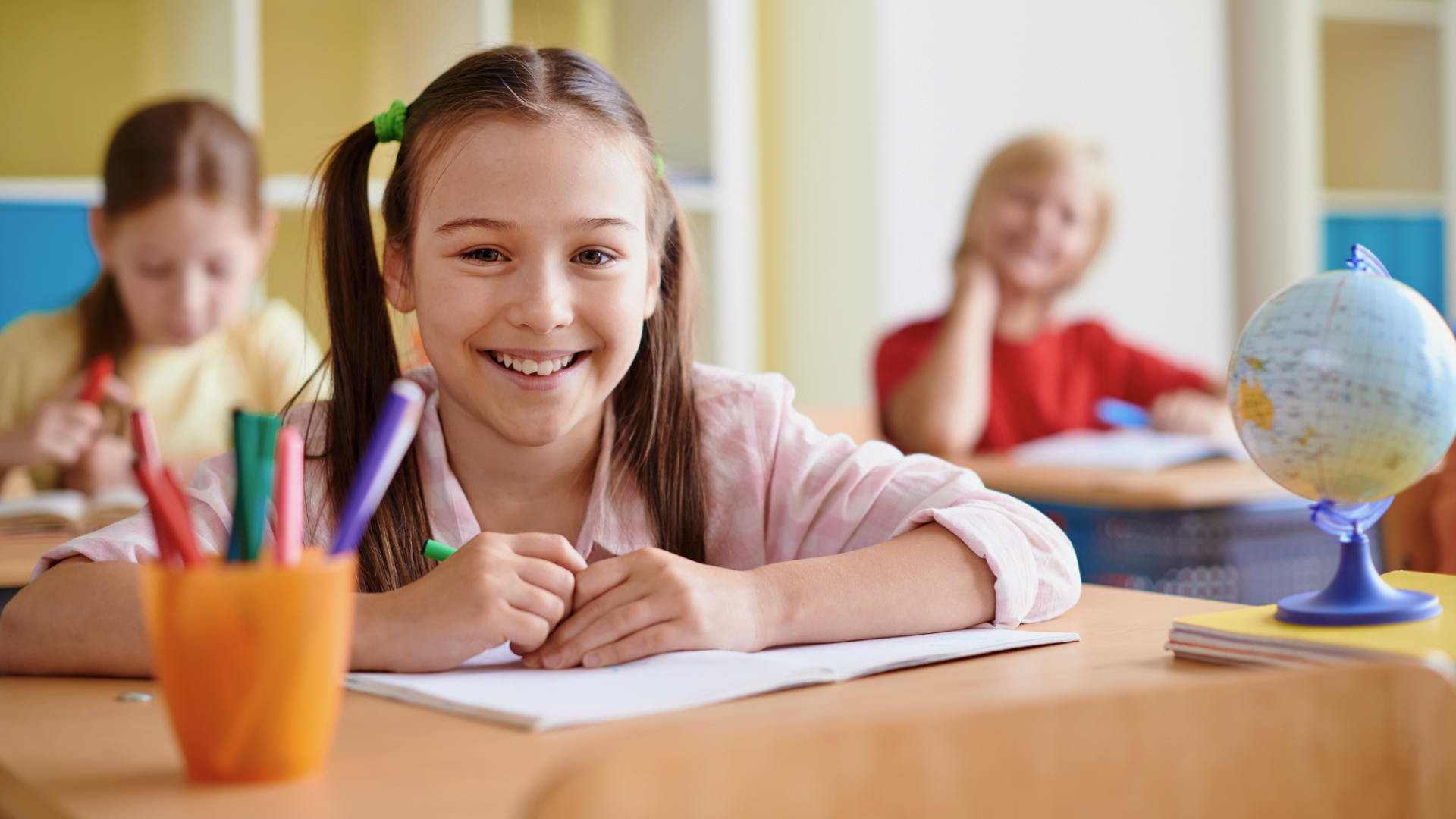 Zdjęcie przedstawia uczniów w klasie szkolnej. Na pierwszym planie znajduje się uśmiechnięta dziewczyna z czymś do pisania w ręku. Przed nią znajduje się otwarty zeszyt, obok niej po prawej stoi globus, a po lewej pojemnik z przyborami do pisania i rysowania. W tle widać dwoje uczniów siedzących w ławkach szkolnych. Zdjęcie w tle jest rozmyte.