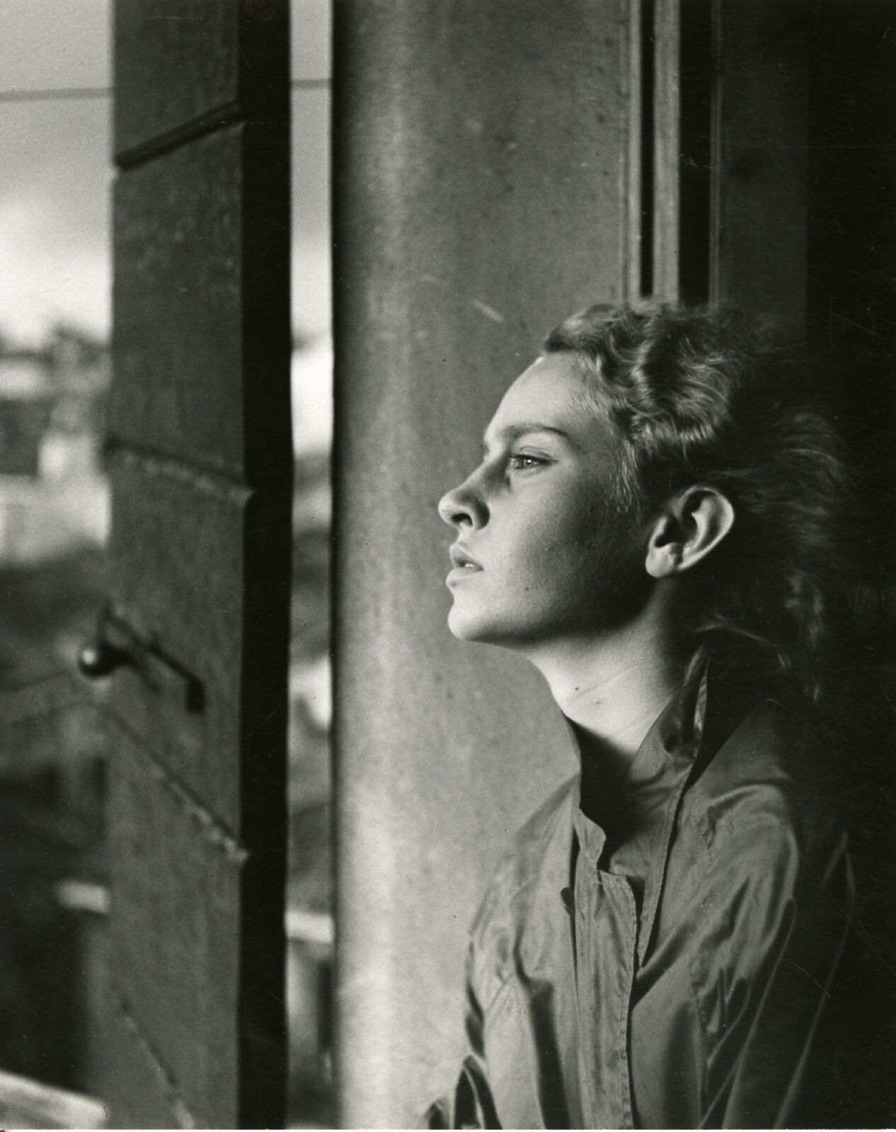 Zdjęcie przedstawia młodą kobietę spoglądającą przed siebie. Uchwycono ją bokiem, od klatki piersiowej w górę. Jej twarz uniesiona jest lekko do góry, oczy delikatnie zmrużone, a usta rozchylone. Jej jasne, kręcone włosy zaczesane są do tyłu. Ma na sobie kurtkę. Twarz kobiety jest najjaśniejszym elementem fotografii. Tło stanowi ciemny front budynku oraz drzwiczki okna, z którego wychyla się bohaterka zdjęcia.