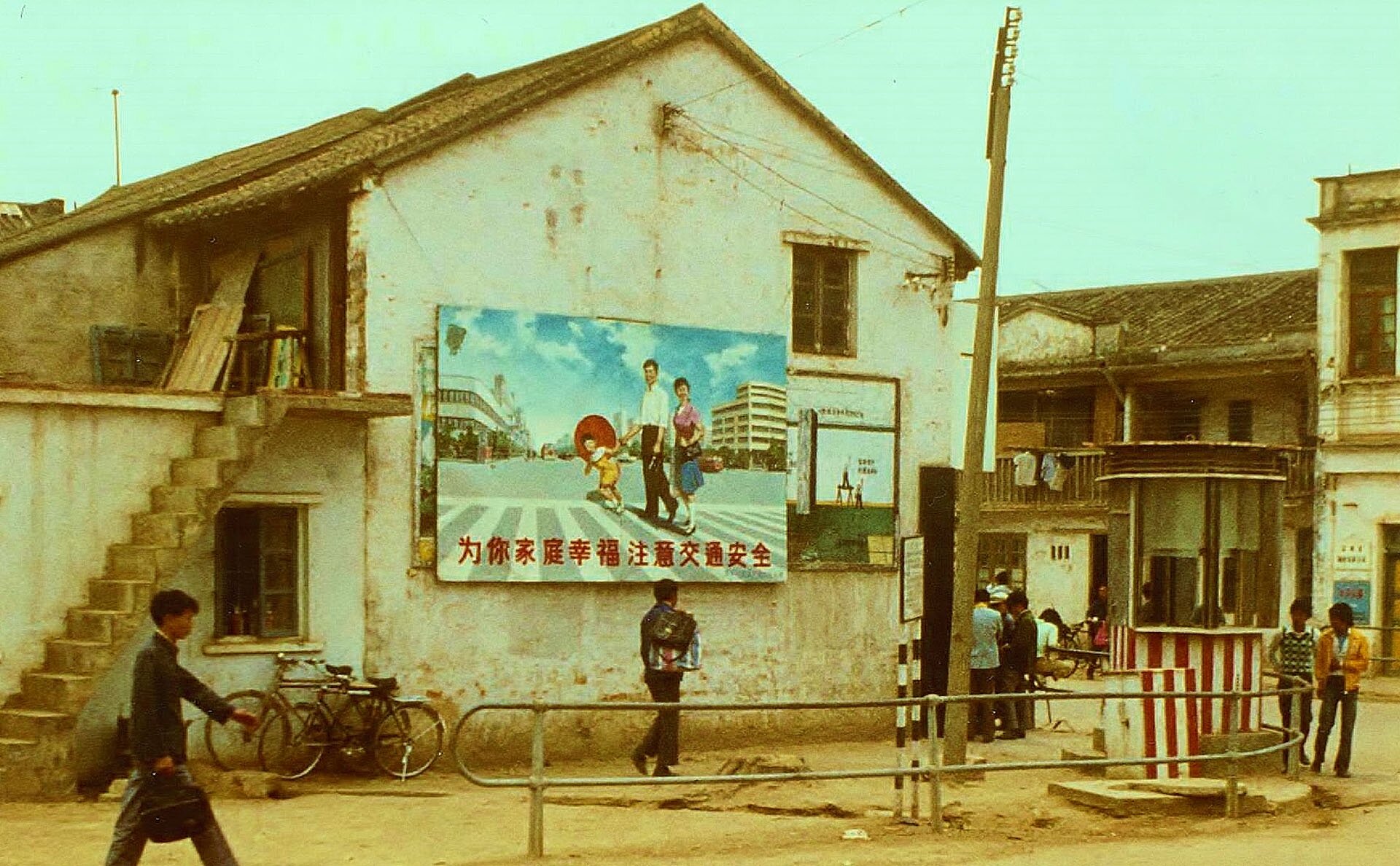 Na zdjęciu ulica, niska zabudowa, na ścianie domu tablica propagandowa. Uśmiechnięta rodzina z jednym dzieckiem.