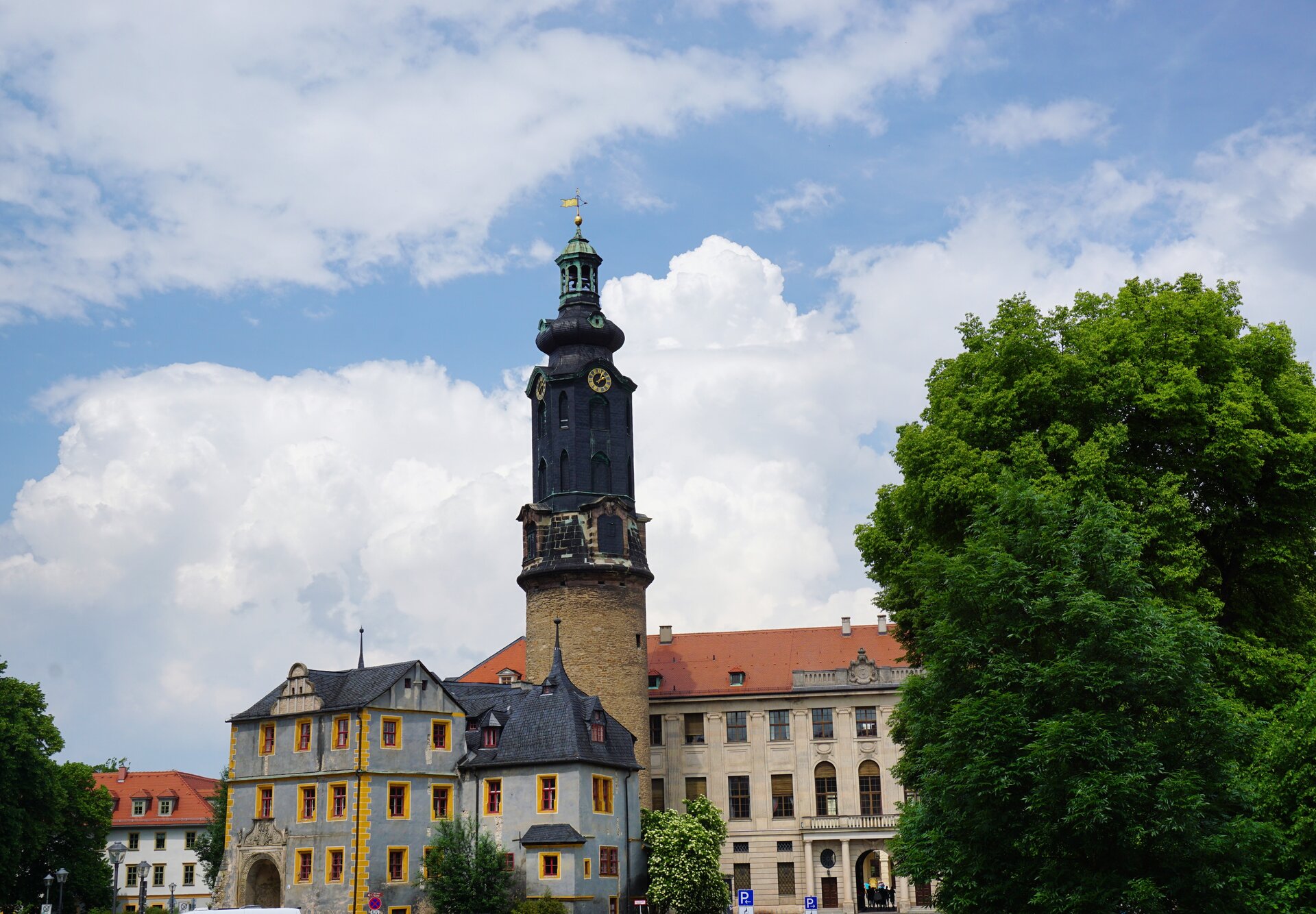 Ilustracja przedstawia wieżę zamkową w mieście Waimar w Niemczech. Wieża zajmuje centralne miejsce na fotografii. Oprócz tego widać jeszcze dwa budynki, szary z żółtymi obramowaniami na oknach i drugi, w kolorze kremowym. Prawą część fotografi zajmuje duże zielone drzewo.