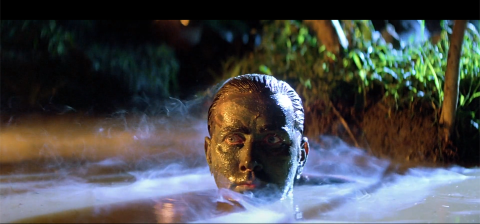 Kadr przedstawia zanurzonego w wodzie mężczyznę. Ukazana jest jego głowa. Mężczyzna ma twarz oraz włosy pomalowane na kolor ciemnozielony. Wokół niego unosi się dym. Patrzy przed siebie. W tle znajduje się roślinność. Panuje noc.