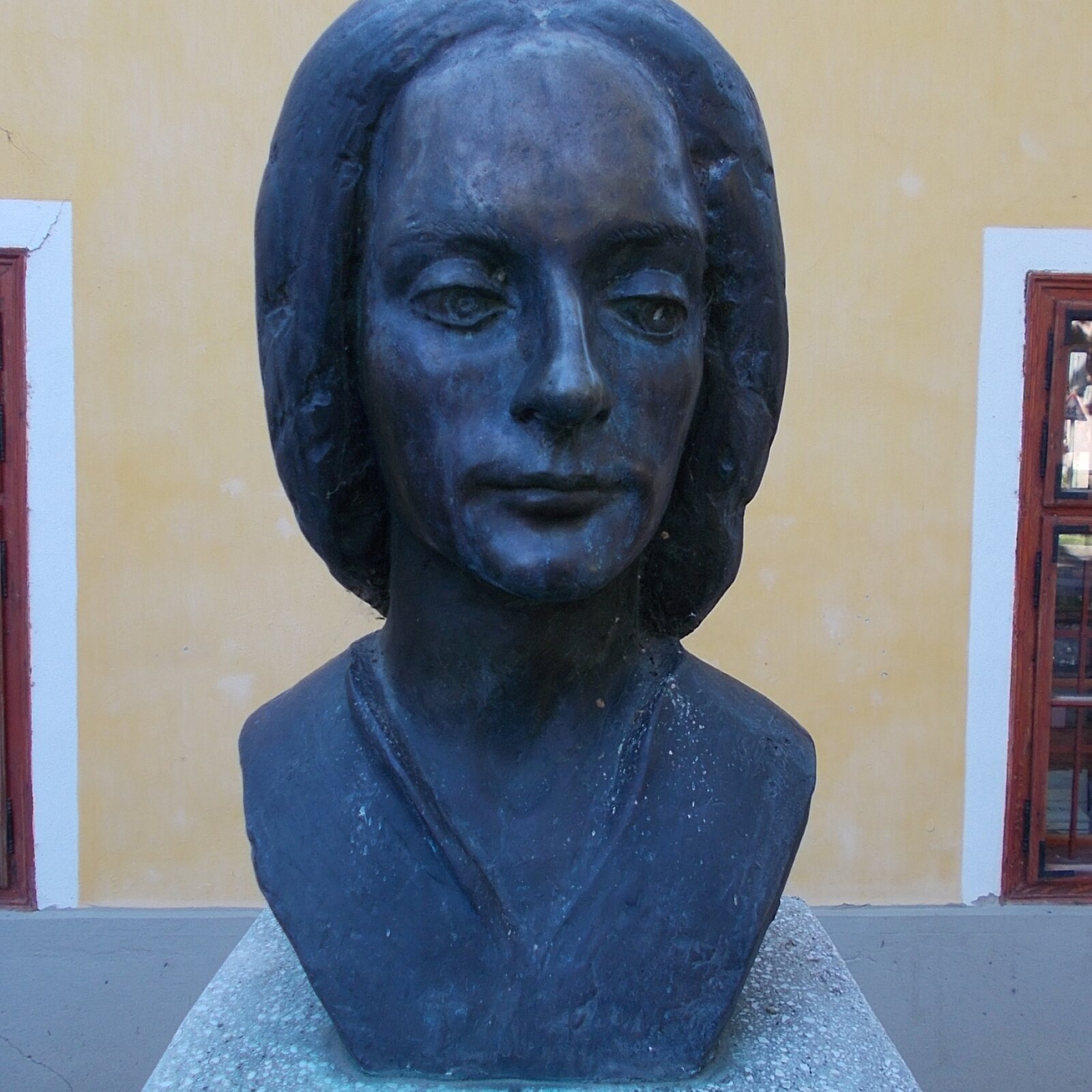 Zdjęcie przedstawia popiersie Antonio Vivaldiego. Popiersie jest wykonane z czarnego materiału i stoi na kamiennej podstawie. Mężczyzna ma gładką twarz, zamknięte usta. Wzrok ma skierowany w dół. Ma grube włosy, zaczesane do tyłu.