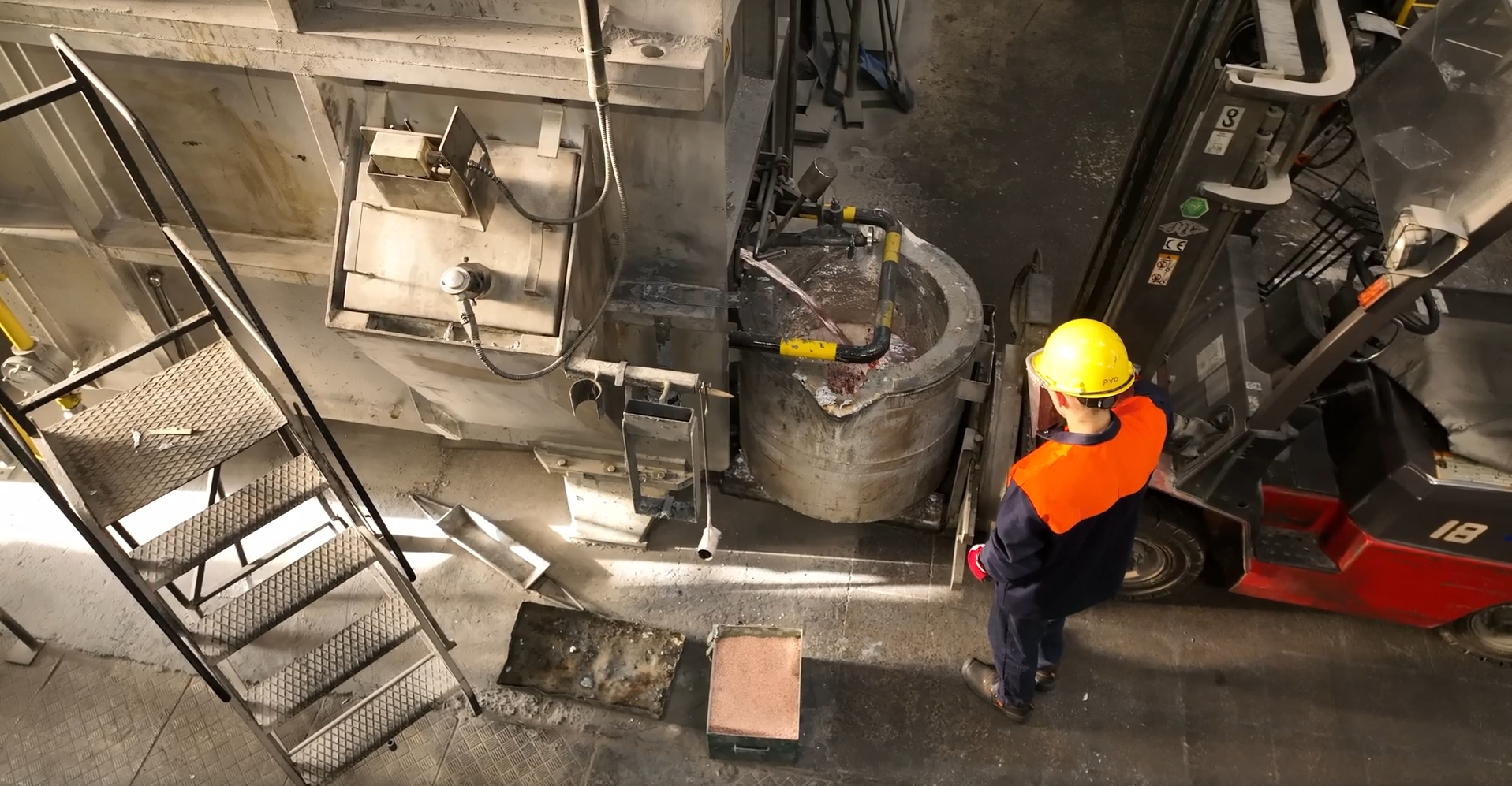 Na zdjęciu widoczna jest przelewanie stopu aluminium do wygrzanej kadzi. W centralnej części zdjęcia znajduje się kadź. Kadź jest w kształcie cylindra. Cylinder jest pusty w środku. Kadź jest oparta przy ścianie. Do kadzi nalewana jest stop aluminium. Procesowi przygląda się pracownik. Pracownik ubrany jest w strój roboczy i kask. Obok pracownika znajduje się wózek widłowy. 