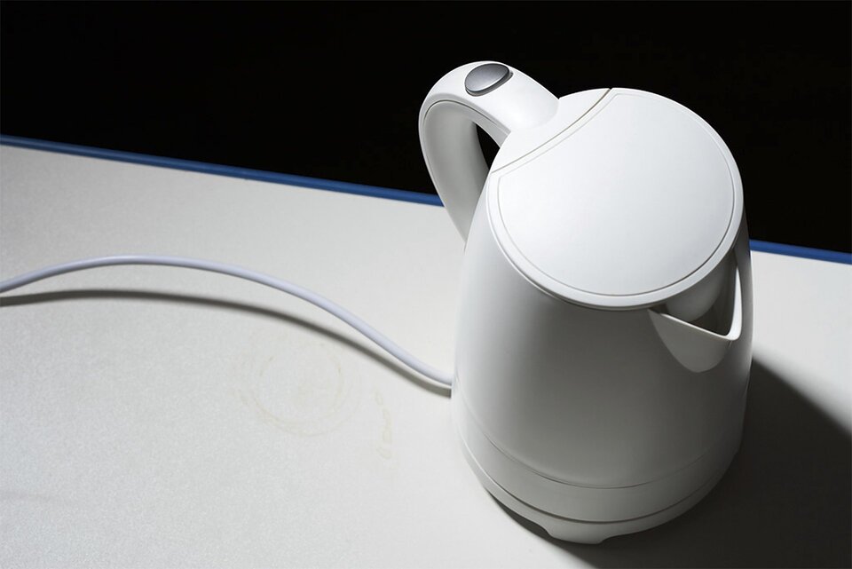 Zdjęcie przedstawia biały czajnik elektryczny stojący na białym blacie kuchennym uchwycony od góry. Czajnik stoi z prawej strony kadru, a oświetlony jest z lewej strony. Tło za blatem czarne.