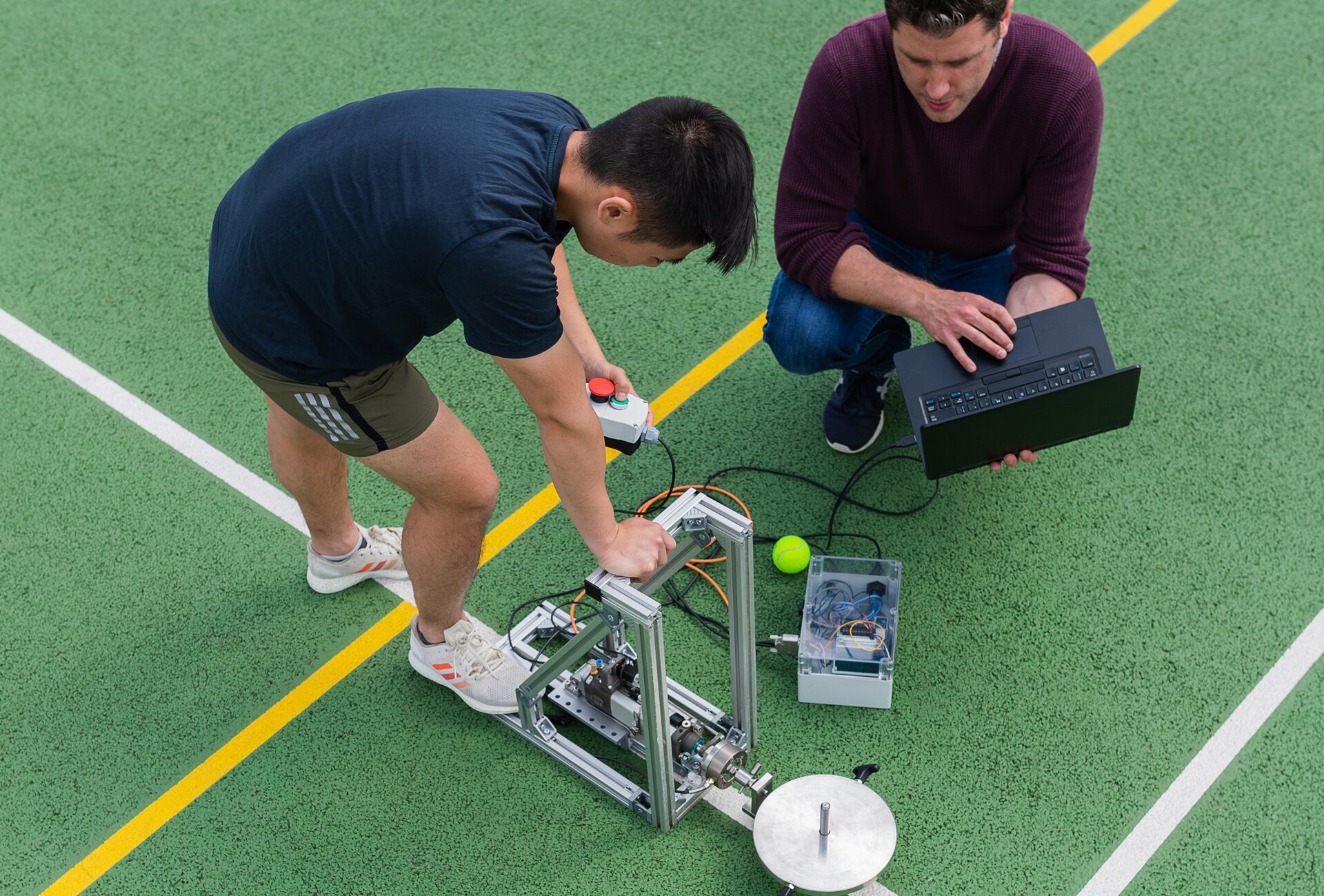 Zdjęcie przedstawia dwie osoby znajdujące się na boisku szkolnym. Widoczny jest mężczyzna w średnim wieku, kucający i trzymający w dłoniach laptopa. Obok stoi chłopiec z urządzeniem pomiarowym, do którego podpięty jest laptop.