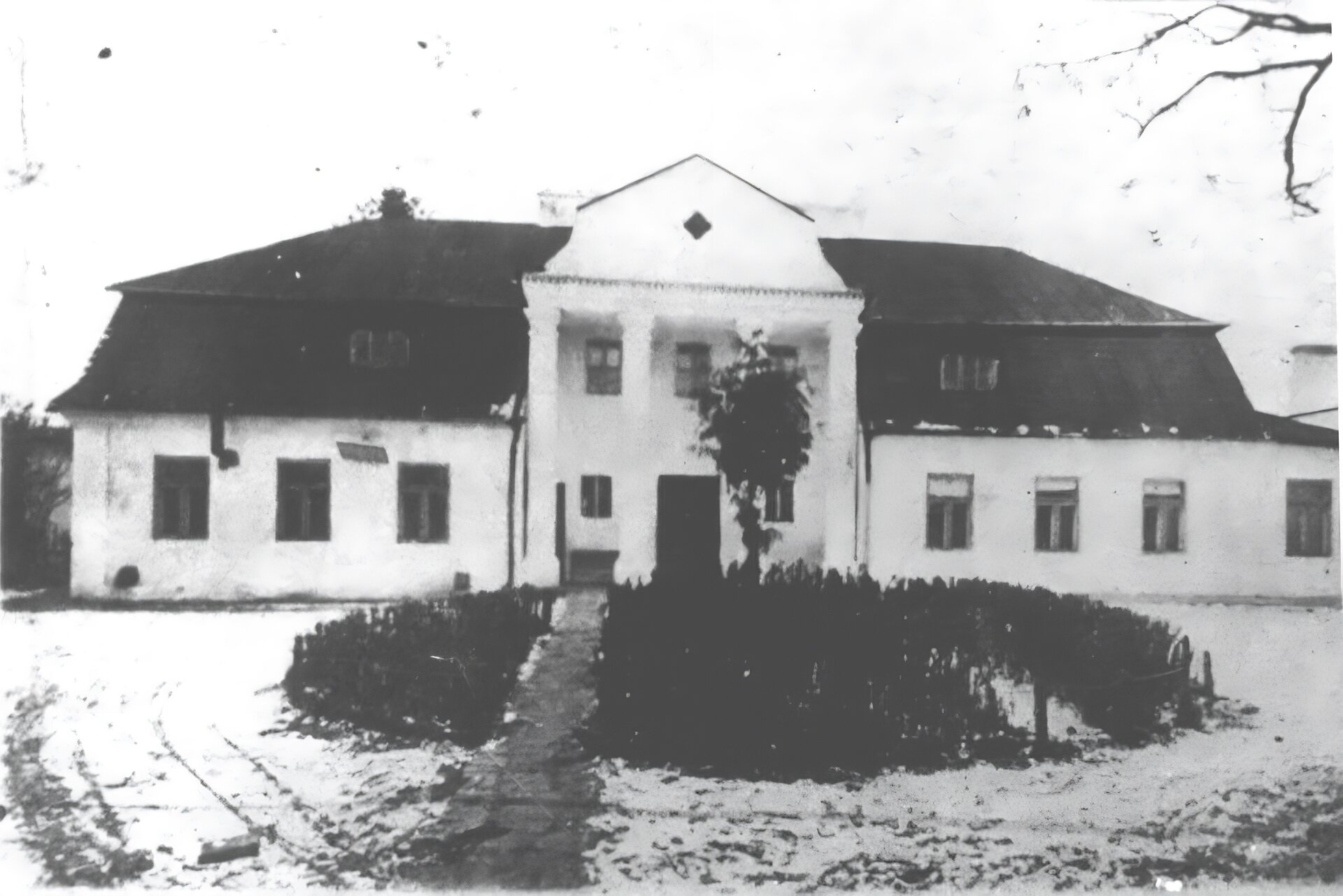 Zdjęcie przedstawia biały, piętrowy dworek. Dach jest skośny i częściowo wysunięty nad wejściem. Podpierają ją cztery kolumny. Przed dworkiem rosną niskie krzewy. Teren wokół dworku jest przykryty śniegiem. Niebo jest bezchmurne. Zdjęcie jest czarno‑białe.