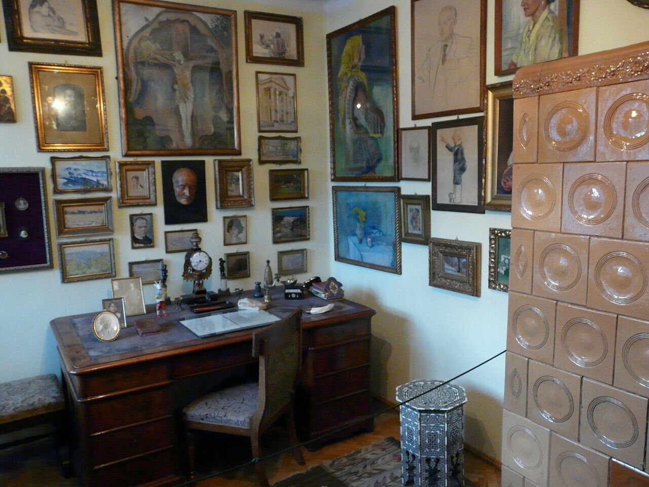 Zdjęcie przedstawia wnętrze pokoju. Znajduje się w nim biurko z krzesłem, duży piec kaflowy i wiele obrazów na ścianach.