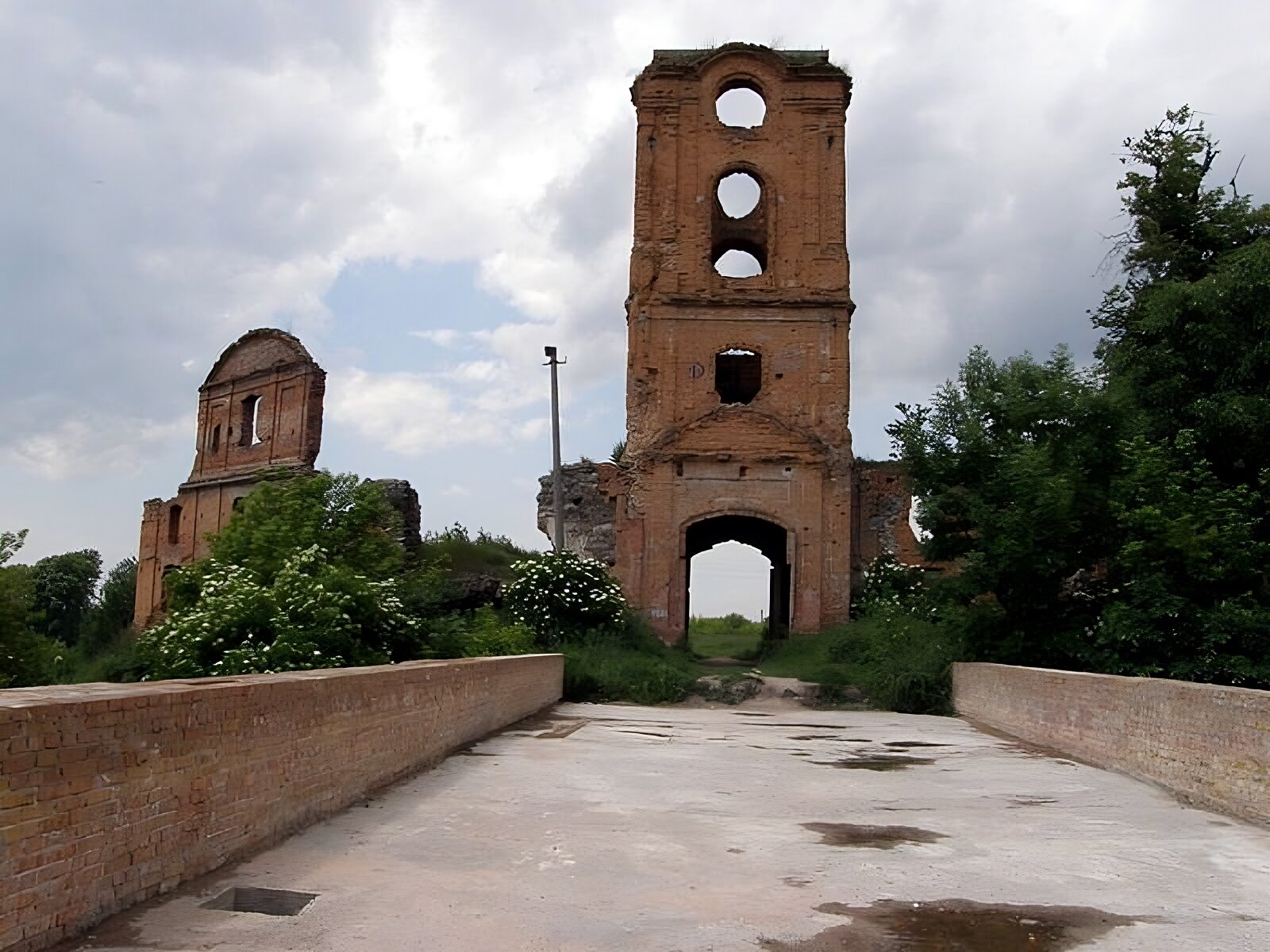 Zdjęcie przedstawia ruiny zamku w Korcu widziane od strony drogi dojazdowej, biegnącej przez murowany most. W centrum fotografii jest fragment trzykondygnacyjnej wieży bramnej, ocienionej pilastrami z tympanonem nad wejściem. Z lewej strony jest zachowany fragment fasady wschodniej, podzielonej pilastrami na części.  Obiekt otoczony jest zielenią. 