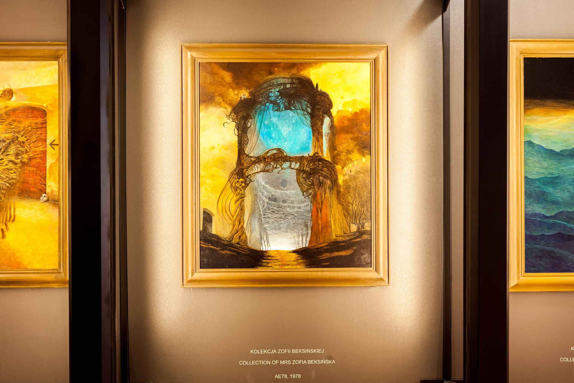 Zdjęcie przedstawia zbliżenie na obraz przedstawiający niebieską przestrzeń, która otoczona jest wieżą z elementów przypominających gałęzie drzewa. Obramowany jest złotymi ramami. Obok znajdują się inne dzieła.