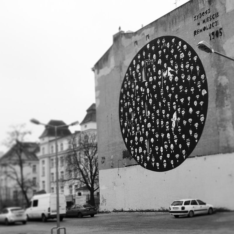 Zdjęcie przedstawia mural na ścianie kamienicy. Mural to koło, w którym znajdują sylwetki różnych ludzi: mężczyzn, kobiet oraz dzieci. Przedstawiają różne grupy społeczne. Na muralu znajduje się flaga PPS, flaga z krzyżem, orłem oraz z napisem po hebrajsku. Nad muralem jest napis: Żyjesz w mieście rewolucji 1905.
