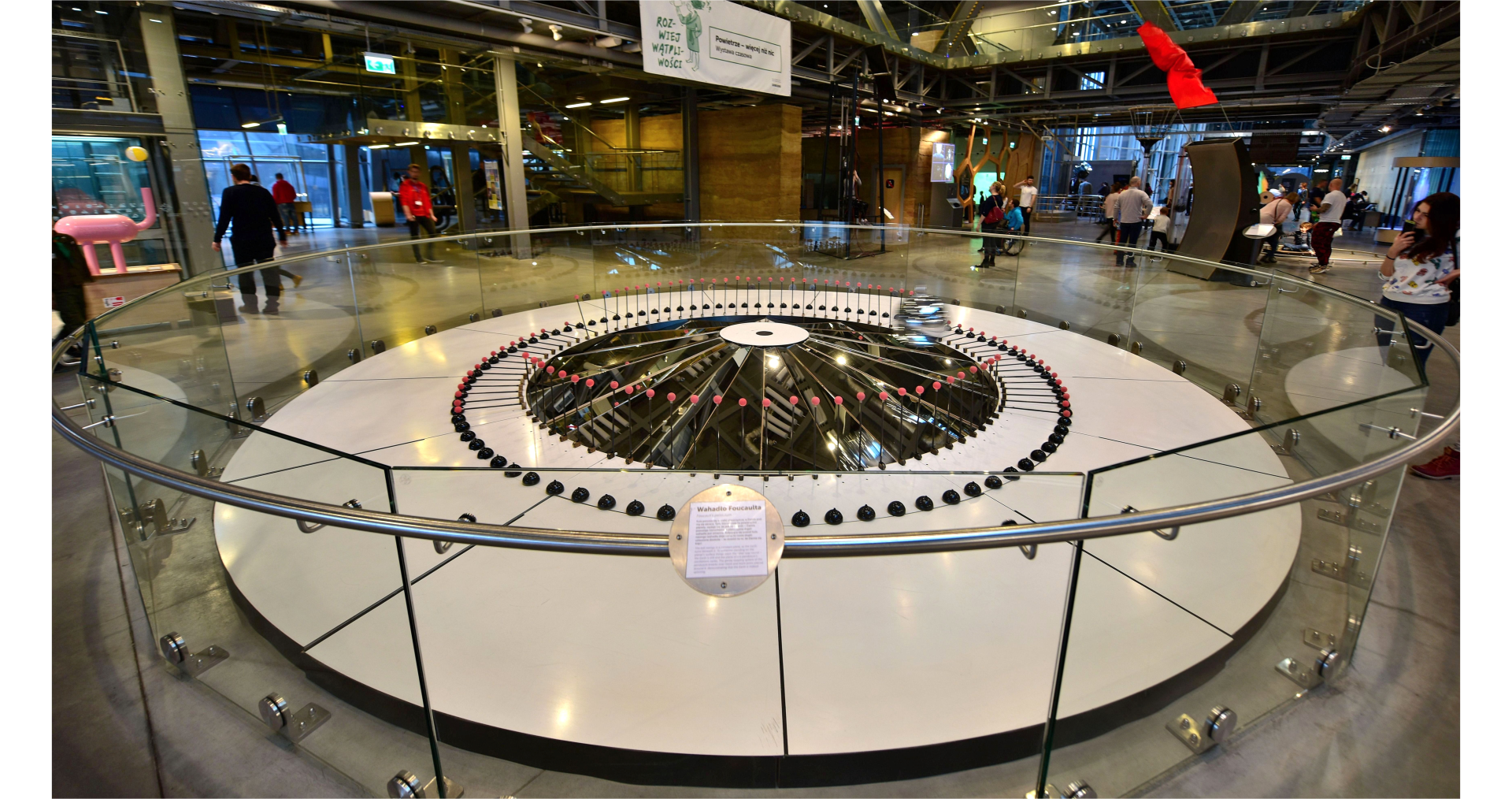 Zdjęcie przedstawia wnętrze wysokiego budynku. We wnętrzu budynku znajduje się ogrodzona strefa, wewnątrz której waha się wahadło Foucaulta. Wahadło strąca ustawione w okrąg pręty, dzięki czemu widać, że nie waha się ono w jednej płaszczyźnie. Wokół strefy małe figurki ludzi.