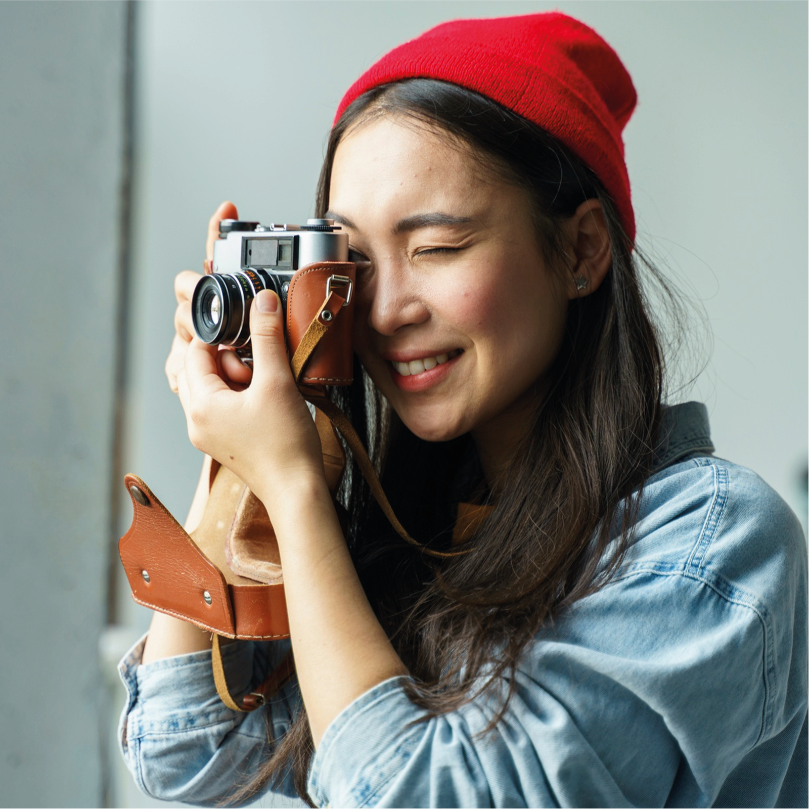 Zdjęcie przedstawia kobietę w czerwonej czapce i dżinsowej kurtce. Jedną z dłoni trzyma aparat, a drugą ustawia ostrość obiektywu. Kobieta uśmiecha się, przymykając jedno oko podczas robienia zdjęcia.