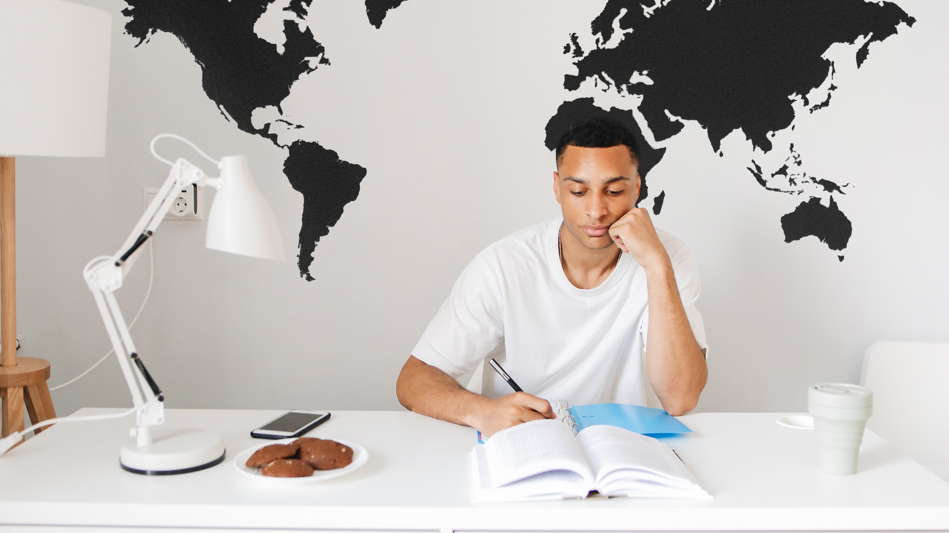 Na zdjęciu chłopak siedzi przy biurku i robi notatki. Na biurku przed nim leży książka i notes, w którym pisze. Po lewej stronie znajdują się lampka, telefon komórkowy i talerz z ciastkami. Po prawej stronie biurka stoi kubek. Za plecami chłopaka, na ścianie jest namalowana mapa świata.