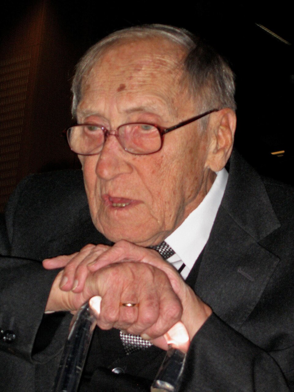 Zdjęcie przedstawia starszego mężczyznę o szczupłej, pociągłej twarzy, ujętego do piersi. Postać ukazuje lewy półprofil. Mężczyzna ma duży nos i wysokie czoło, na nosie druciane okulary, oczy są głęboko osadzone. Ma krótkie, proste, rzadkie włosy z wyraźną łysiną. Ubrany jest w garnitur, koszulę i krawat. Dłonie opiera na lasce. Na palcu prawej ręki ma złotą obrączkę. Tło jest czarne.