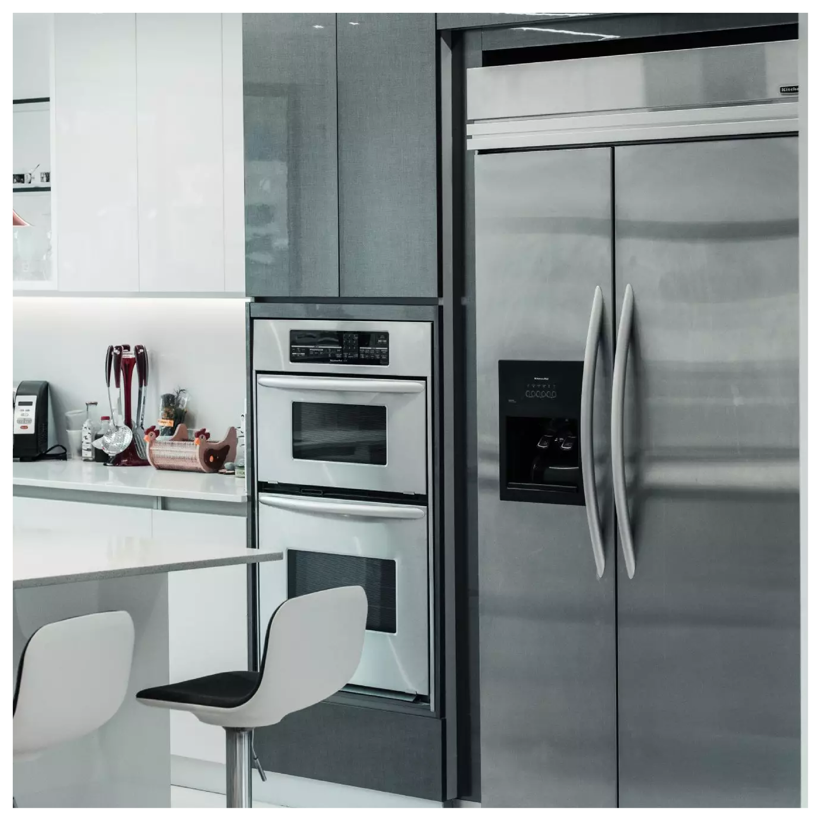 Widoczny jest fragment kuchni. Przy wysokim blacie stoją krzesła. Po prawej stronie piekarnika stoi lodówka. Po lewej stronie piekarnika są szafki i blaty.