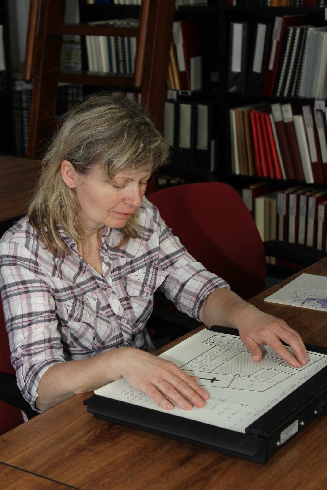 Ilustracja przedstawia kobietę, której dłonie spoczywają na rysunku technicznym, leżącym na czarnym dużym panelu. Rysunek ma wypukłe elementy. Obok widoczny jest tekst zapisany alfabetem Brajla.