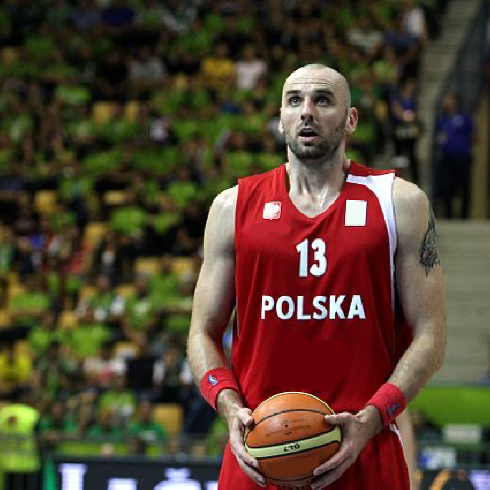 Zdjęcie przedstawia Marcin Gortata w czerwonej koszulce bez rękawów, z piłką do koszykówki w dłoniach.