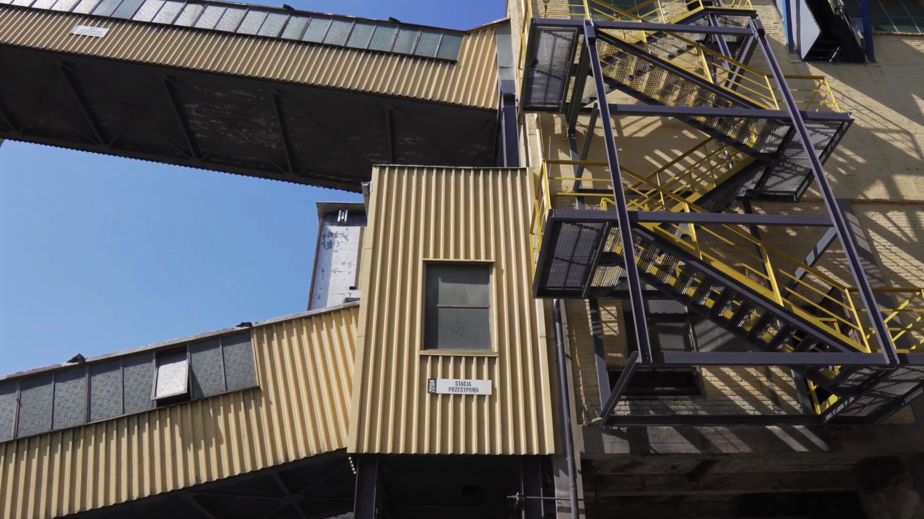 N zdjęciu widać fragment mostu transportowego. W centralnej części ekranu widoczny jest budynek z którego wychodzi łącznik. Z prawej strony obiektu widoczna jest klatka schodowa. Klatka jest zabezpieczona barierką.