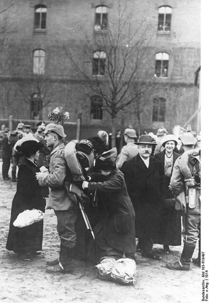 Kliknij żeby powiększyć Na zdjęciu niemieccy żołnierze. Stojący tyłem lub bokiem. Na plecach mają wojskowe plecaki m.in. z kocami. Jedna z osób pakuje coś jeszcze żołnierzowi do plecaka. Żegnający trzymają także paczki, zawiązane sznurkiem. Nastrój dość radosny, zwłaszcza mężczyzna w meloniku i płaszczu i kobieta w kapeluszu patrzący na fotografa, uśmiechają się. Z tyłu widoczne okna budynku, z łukowatymi zakończeniami..