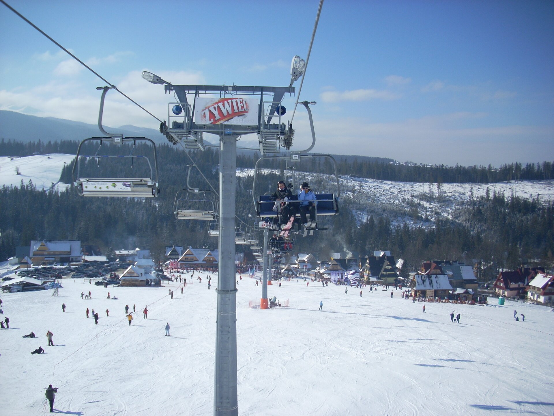 Zdjęcie wykonane zimą na wyciągu narciarskim. Pośrodku fotografii linia słupów, bo obu stronach zamontowane liny, do których przymocowano ławki wyciągu. Siedzą na nich narciarze. W dole rozległy stok narciarski, dziesiątki zjeżdżających osób. Na drugim planie, u podnóża stoku budynki o stromych dachach, a w tle kolejne wzniesienia.