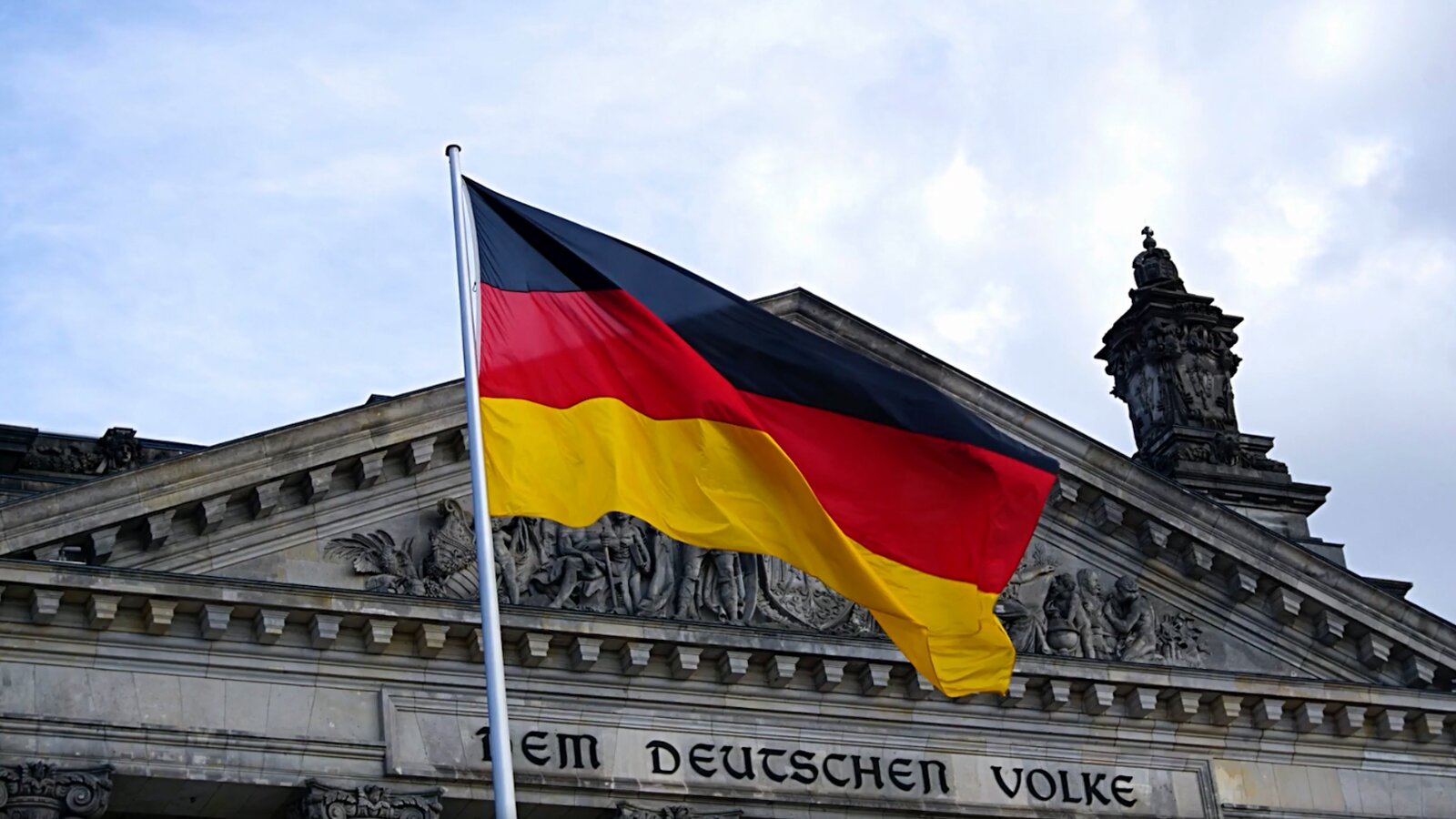   Sprachkenntnisse zu verbessern, ist ein Auslandsaufenthalt. Nur in einem deutschsprachigen Land kommst du ständig in Kontakt mit Native Speakern und sprichst die ganze Zeit Deutsch. Dabei lernst du, den Alltag in dieser Sprache zu bewältigen. Bereits während der Schulzeit gibt es Möglichkeiten, Auslandserfahrung zu sammeln, beispielsweise durch einen Schüleraustausch oder durch ein Auslandsschuljahr. Nach dem Abi hast du zudem die Möglichkeit, dich für ein Studium in Deutschland, Österreich oder in der Schweiz zu entscheiden.
4. Den Alltag vor Ort erleben
Die beste Möglichkeit, deine Sprachkenntnisse zu verbessern, ist ein Auslandsaufenthalt. Nur in einem deutschsprachigen Land kommst du ständig in Kontakt mit Native Speakern und sprichst die ganze Zeit Deutsch. Dabei lernst du, den Alltag in dieser Sprache zu bewältigen.
Opis: Ilustracja ukazuje szczyt budynku niemieckiego parlamentu, na którym widnieje napis   "Dem Deutschen Volke ". Obok budynku powiewa flaga Niemiec, składająca się z trzech poziomych pasów w kolorach czarnym, czerwonym i żółtym.
