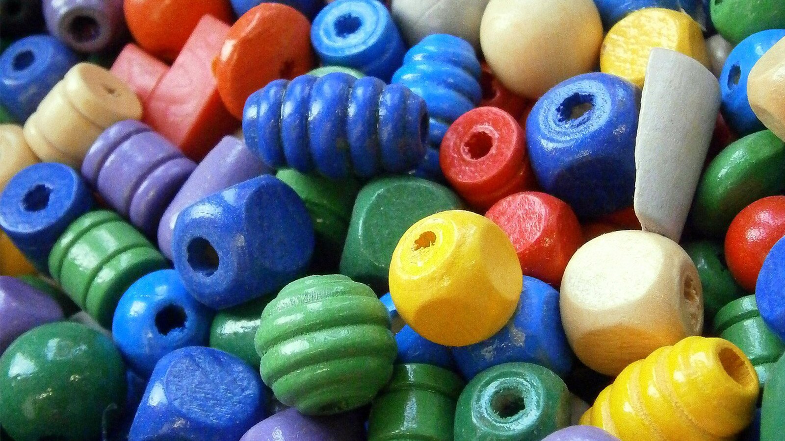 Zdjęcie przedstawia różnokolorowe koraliki, które mają różne kształty. Widoczne są paciorki prostopadłościenne, kuliste i owalne z żeberkowaniem, walcowate i w kształcie dysku. Paciorki mają kolory: niebieski, zielony, żółty, czerwony i kremowy.