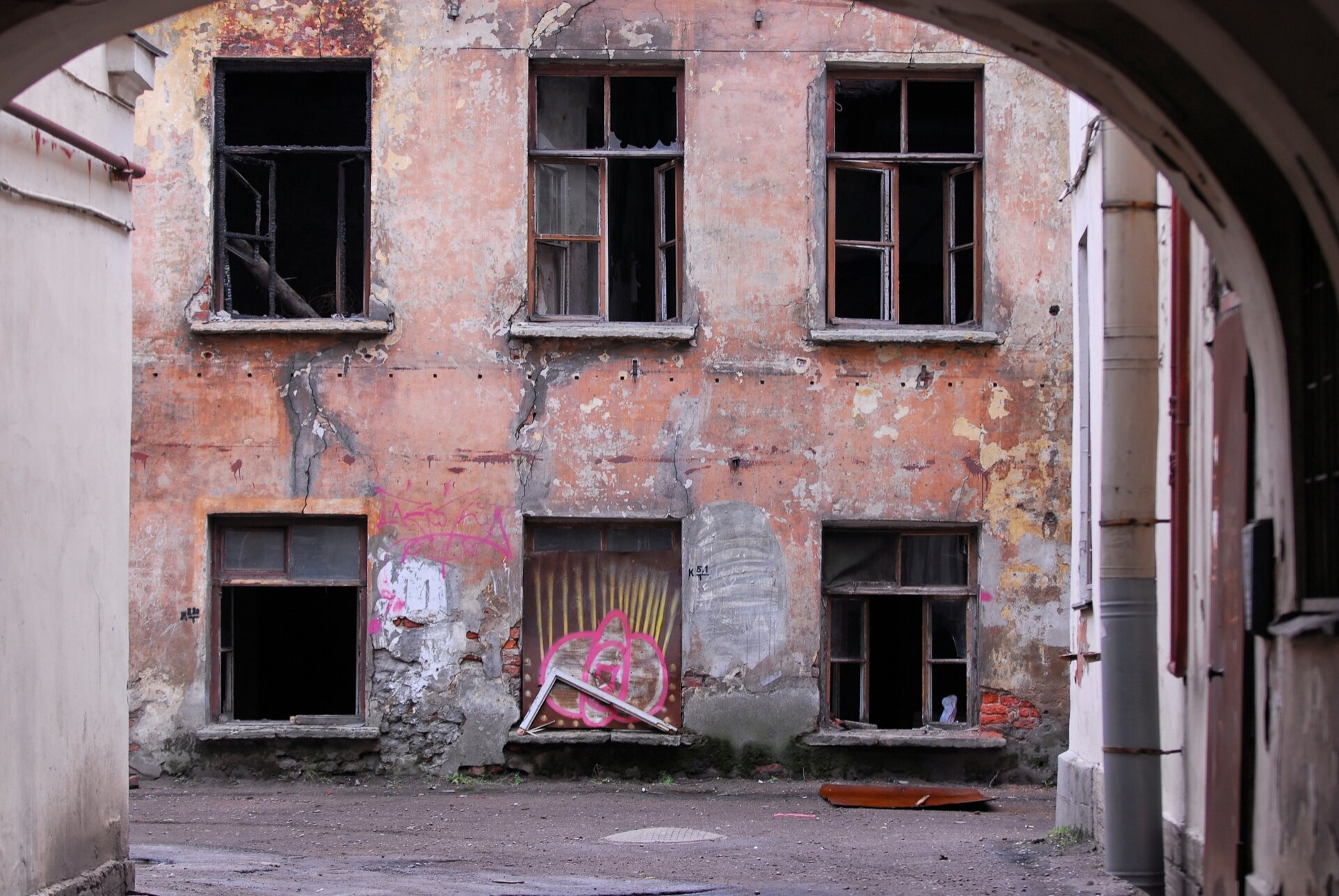 Zdjęcie przedstawia ścianę budynku. Widoczne są wybite okna oraz obdrapana  ściana. Zdjęcie wykonano z perspektywy półokrągłego wejścia na dziedziniec. 