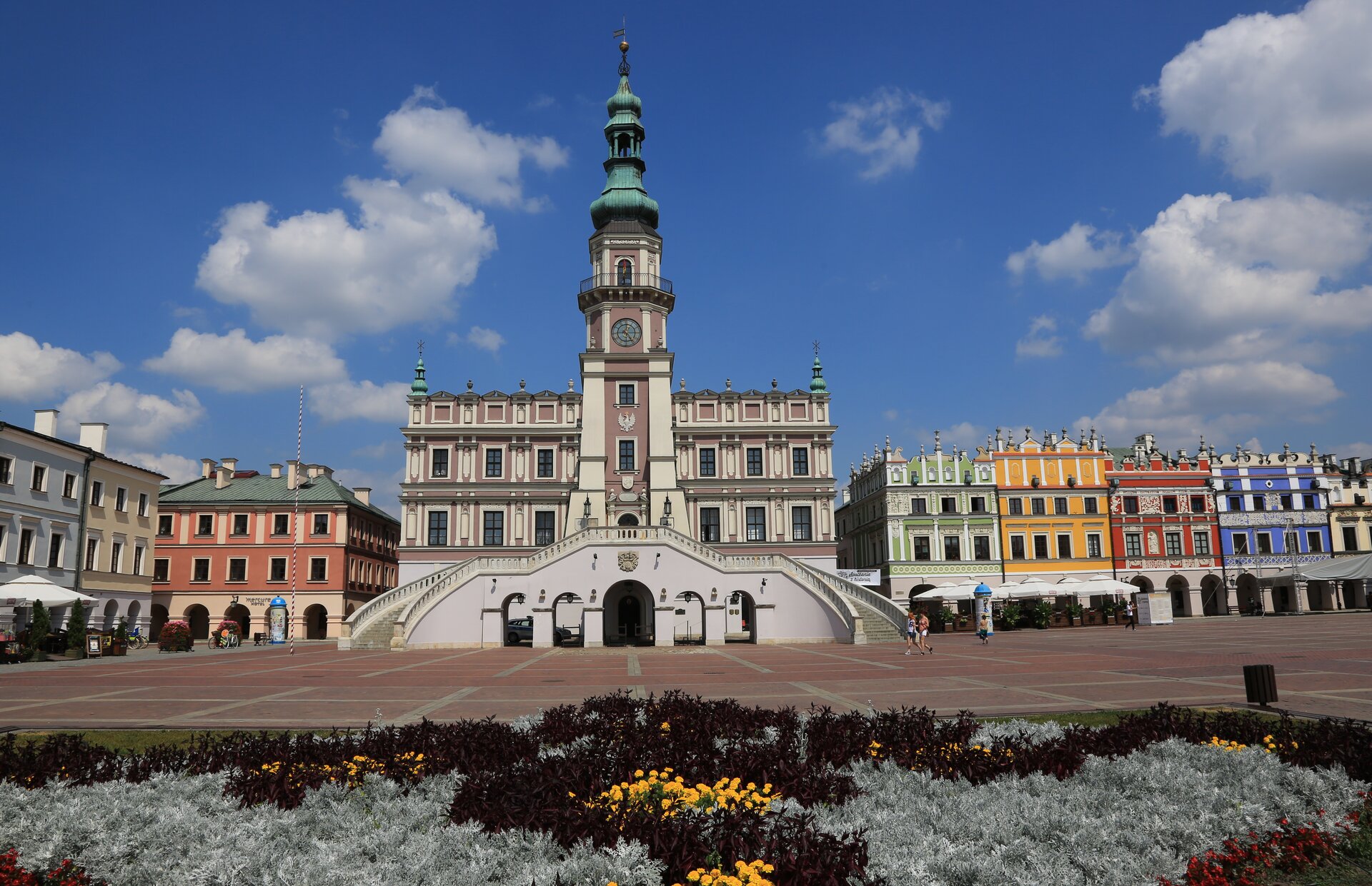 Na zdjęciu ratusz z wysoką wieżą i okazałym schodami. Obok kolorowe, odrestaurowane kamieniczki. Przed ratuszem rozległy plac, kwietnik.