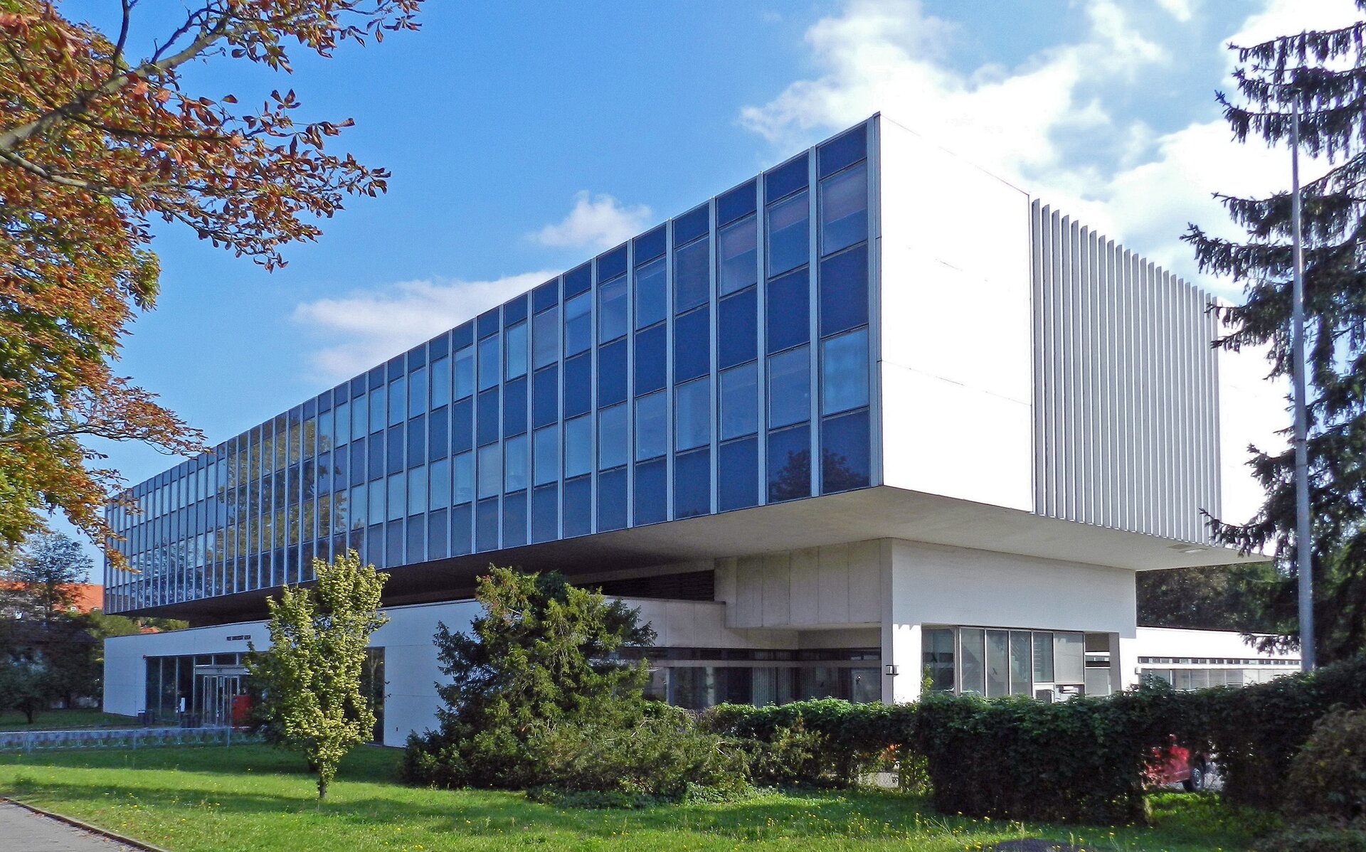 Fotografia przedstawia budynek, przed którym rosną krzewy oraz drzewa. Budynek ma fasadę koloru białego (ściany boczne i dolne) oraz niebieskiego (czoło budynku). Okna znajdują się w niebieskiej części fasady, w dwóch rzędach. Poniżej okien znajduje się wejście do budynku. Po prawej stronie budynku - parking, na którym stoi czerwone auto.