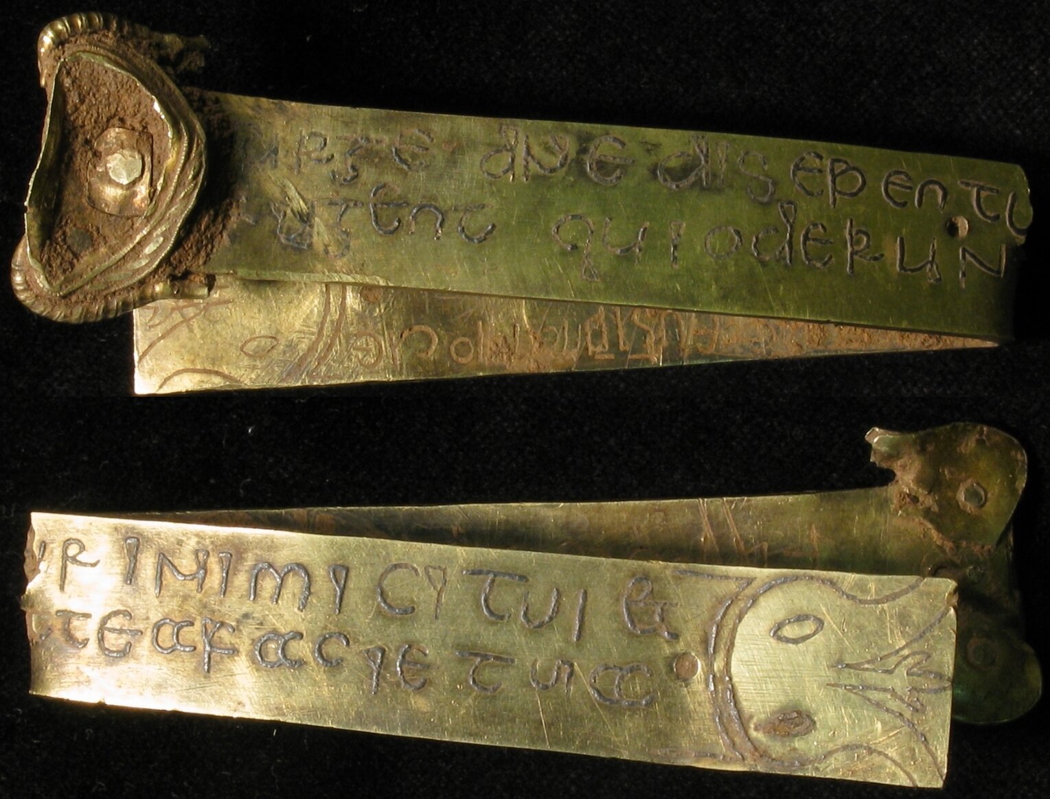 Metalowy pas z fragmentem zapięcia, którego zdjęcie wykonano podwójnie, aby przedstawić obie jego strony. Zdjęcie współczesne eksponatu, wykonane na czarnym tle. Widoczny zapis tekstu łacińskiego w dwóch wersach. 
