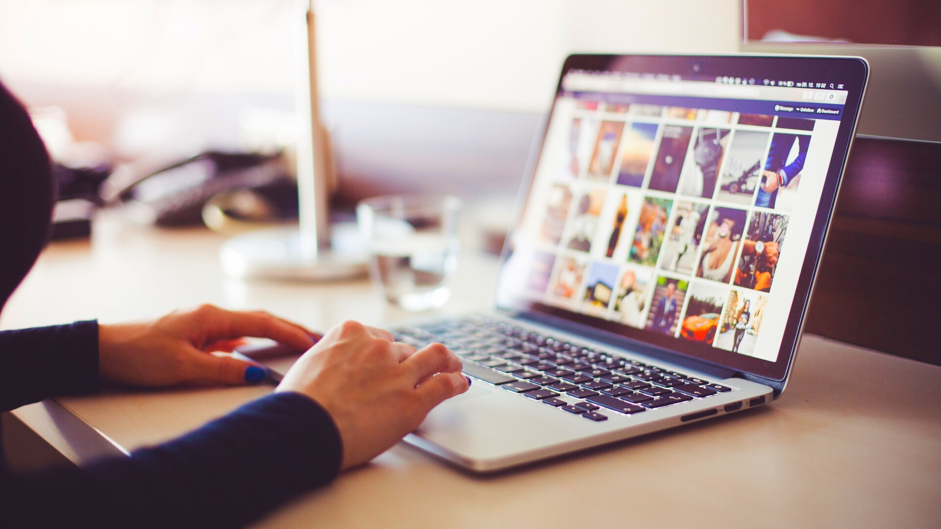 Ilustracja przedstawia laptop leżący na biurku. Na klawiaturze widać kobiece dłonie. Monitor przedstawia różnego rodzaju grafiki. Prawdopodobnie kobieta szuka odpowiedniego obrazu do swojego projektu. W tle widać szklankę wody, a jeszcze dalej telefon. Kobieta być może znajduje się w biurze.