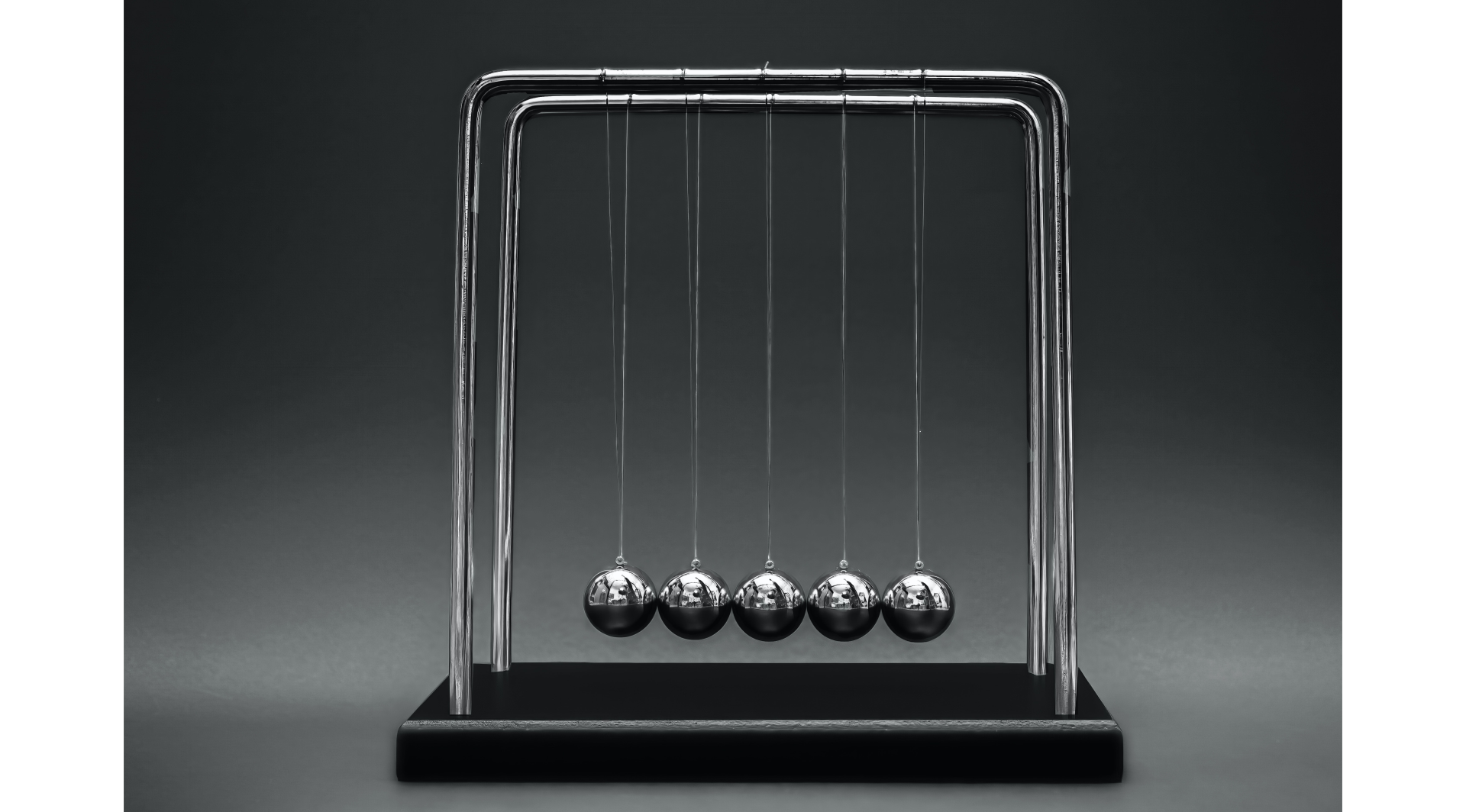 Zdjęcie. Tło szare. W centralnej części stojak na którym zawieszonych jest pięć wahadeł. Stojak ma dwa ramiona, a kulki wahadeł stykają się i są zawieszone równolegle na niciach zaczepionych na każdym z ramion stojaka. Kulki są metalowe, widać w nich odbicie osoby robiącej zdjęcie.