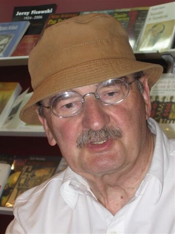 Zdjęcie przedstawia starszego mężczyznę. Ma prostokątną twarz ze zmarszczkami, wydatnym nosem oraz siwymi wąsami. Ma założone okulary w owalnych oprawkach oraz brązowy kapelusz na głowie. Ubrany jest w białą koszulę. Za mężczyzną znajduje się półka z książkami.