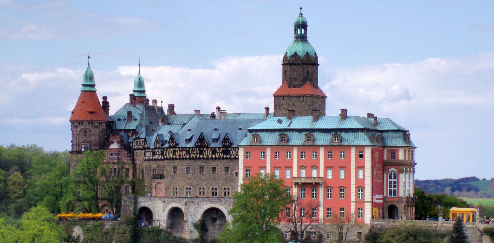 Na zdjęciu okazały zamek o niejednolitej budowie, z przodu i po lewej stronie otoczony drzewami. Umiejscowiony jest na wysokim, urwistym, skalnym zboczu, więc góruje nad drzewami.  Z lewej strony, na krótszym boku budowli, widoczne są dwie baszty, na samym dole - taras ze stolikami i kilkoma rozłożonymi parasolami. Środkowa część zamku u podstawy ma arkady z trzema szerokimi otworami zakończonymi łukami. Do wysokości kolejnych dwóch pięter ściana pozbawiona jest elementów ozdobnych, widać zamurowane łukowate wejście, powyżej niego mały balkon, poza tym dwa rzędy prostych okien. Poziom wyżej rozpoczyna się poddasze, którego elewacja jest otynkowana na jasny kolor i ozdobiona elementami drewnianymi. Dach w całości pokryty jest blachą, wkomponowano w niego liczne wykusze, lukarny, okna tzw. wole oczy oraz kominy. Nieco na prawo widoczna jest potężna, czworoboczna wieża, przylegająca do trzypiętrowego budynku, który sprawia wrażenie zbudowanego w późniejszym okresie. Jest otynkowany na jaskrawy kolor, ma też jasne pionowe elementy ozdobne, okna są białe. Na dole po prawej stronie jest kolejny taras lub dziedziniec zabezpieczony kamienną balustradą. Stoi na nim rozkładany pawilon, widać grupę ludzi.