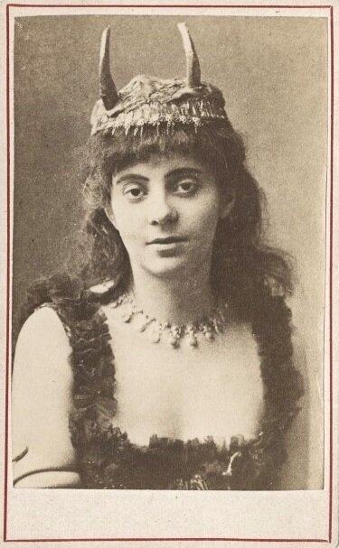 Stara fotografia przedstawia portret młodej kobiety o łagodnym wyrazie twarzy. Kobieta ma ciemne, długie włosy opadające do ramion oraz nakrycie głowy z dwoma rogami. Na szyi bogaty naszyjnik oraz duży dekolt.