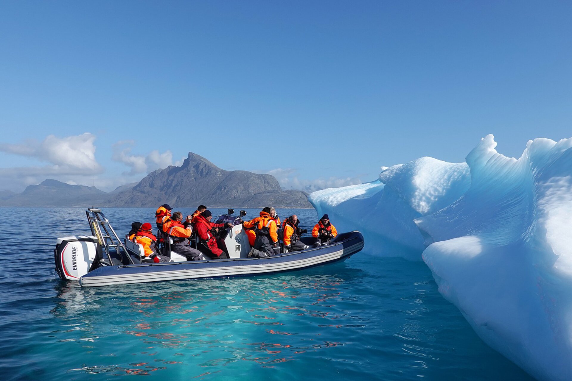 Fotografia przedstawia ludzi w skafandrach na motorówce, podpływających do góry lodowej. W tle skalista góra i jasne niebo.