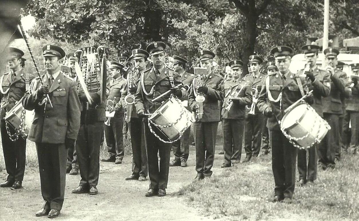 Zdjęcie przedstawia orkiestrę wojskową grającą w plenerze. Mężczyźni stoją w czterech rzędach. Przed nimi kapelmistrz.