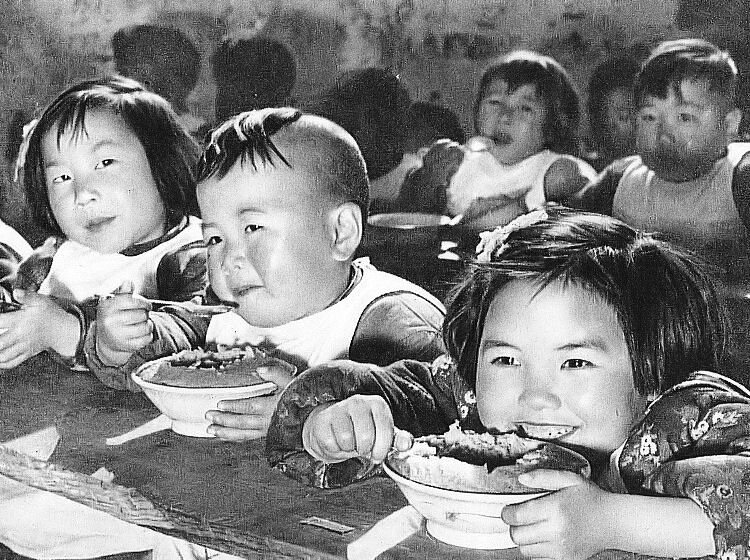 Fotografia przedstawia azjatyckie małe dzieci, chłopców i dziewczynki, siedzących obok siebie i spożywających posiłek. Dzieci uśmiechają się.