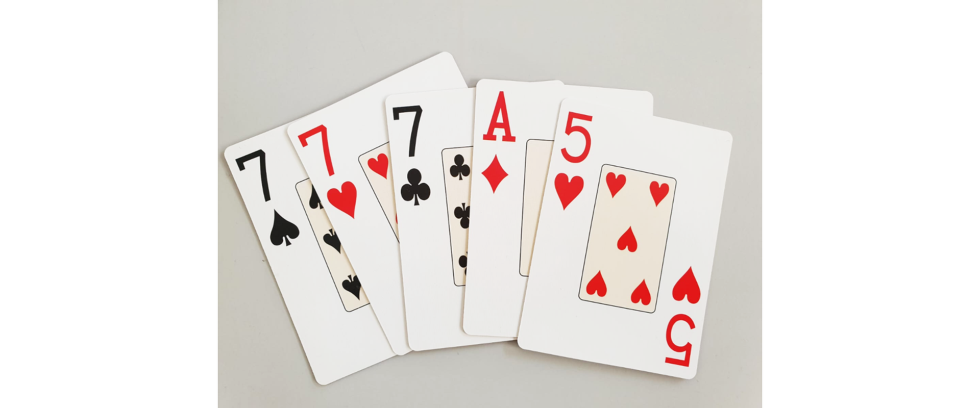 Ilustracja przedstawia pięć kart wyłożonych na szarym tle. Karty od lewej to siódemka pik, siódemka kier, siódemka trefl, As karo, piątka kier.