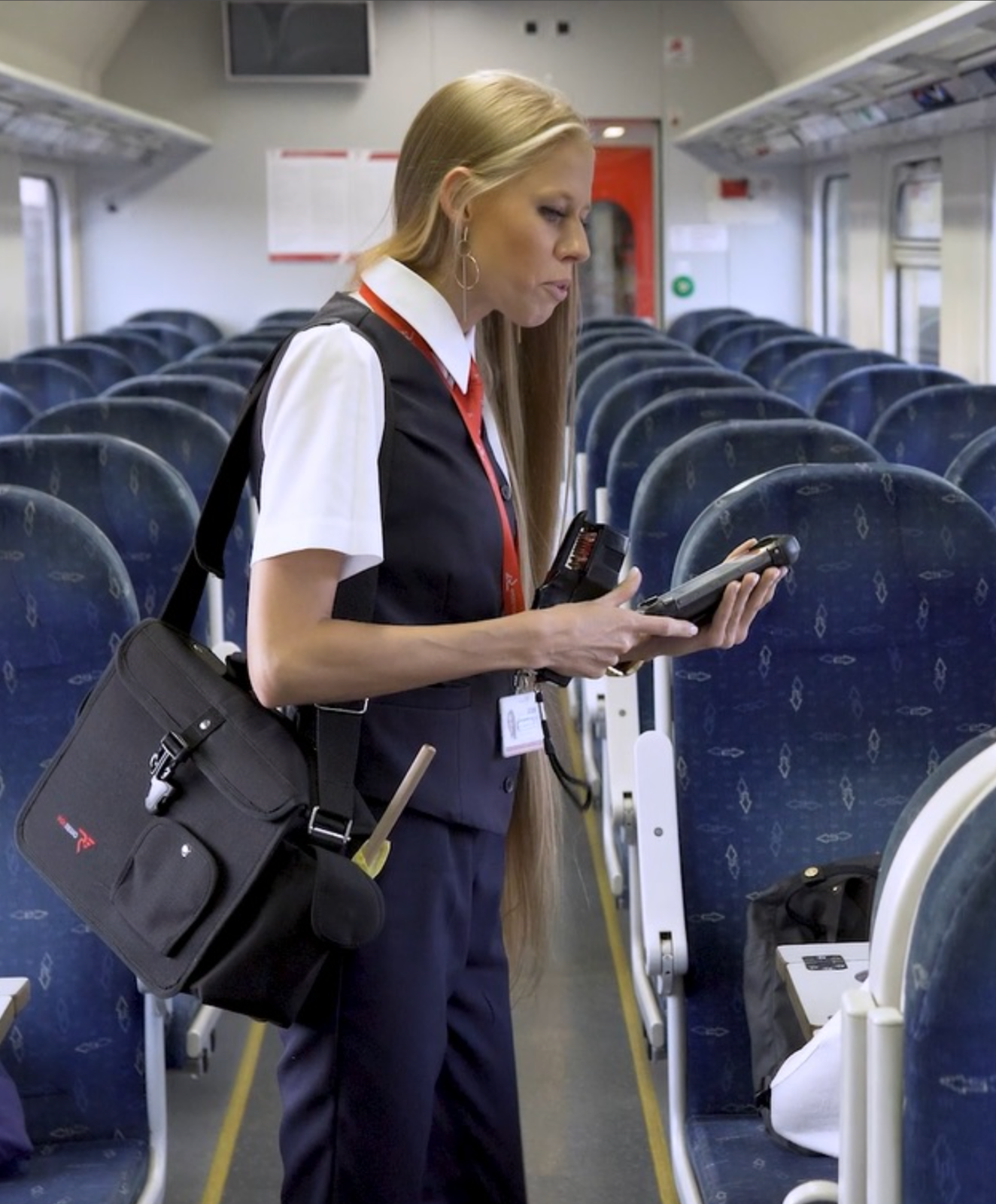 Zdjęcie przedstawia konduktora. Na zdjęciu widać kobietę stojącą między fotelami wagonu pasażerskiego. Trzyma w rękach czytnik biletowy.