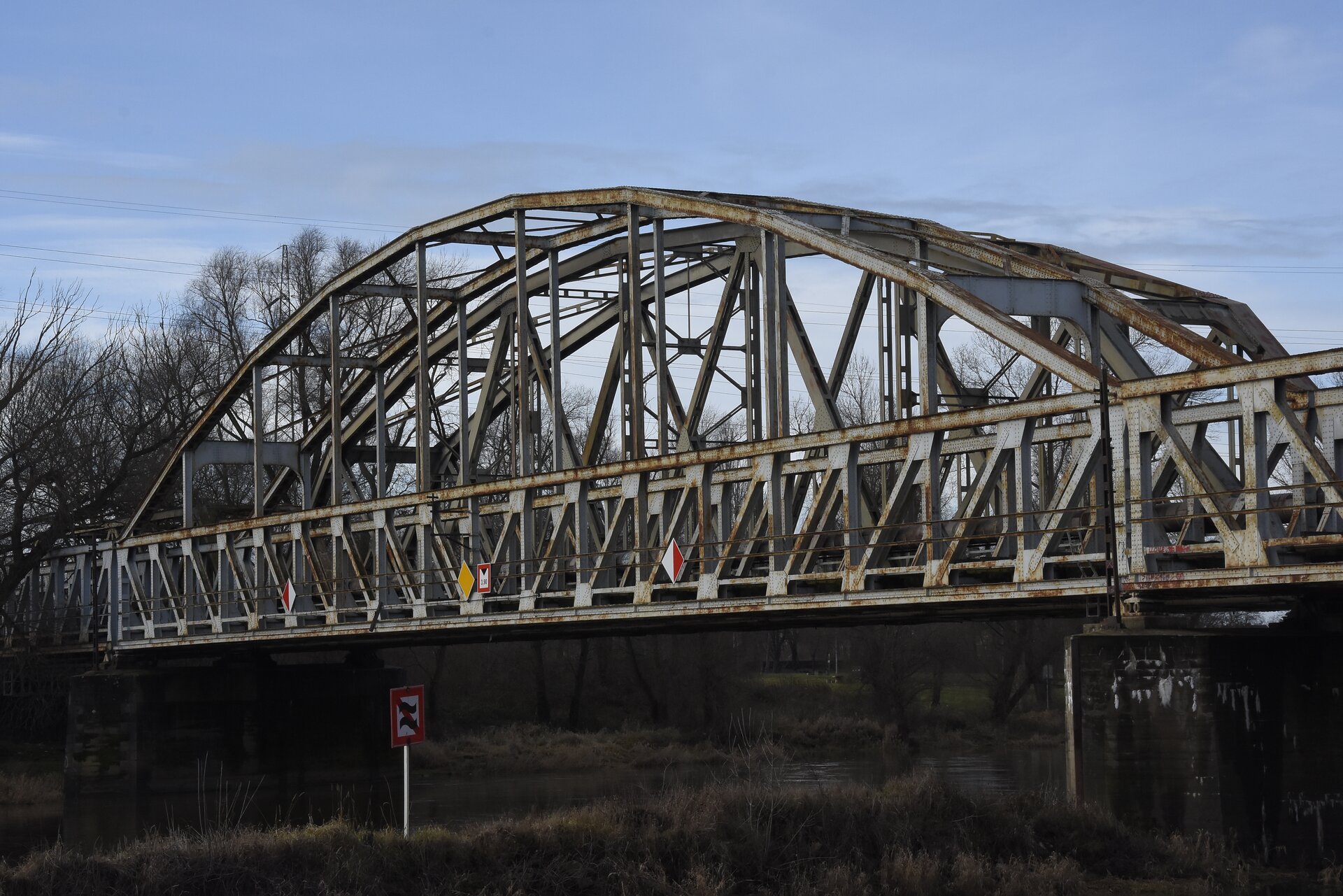 Ilustracja przedstawia most o konstrukcji łukowej, w którym powierzchnia jezdna jest usytuowana u dołu. Most jest konstrukcją stalową wspartą na żelbetowych podporach. Zarówno konstrukcja łuku, jak i kładki jezdnej oraz barierek, zostały pomalowane na kolor biały, przy czym na niektórych elementach widoczne są ślady rdzy. Ujęcie przedstawia most z boku, co pozwala dobrze widzieć żelbetowe podpory oraz łożyska znajdujące się pomiędzy podporami a belkami.