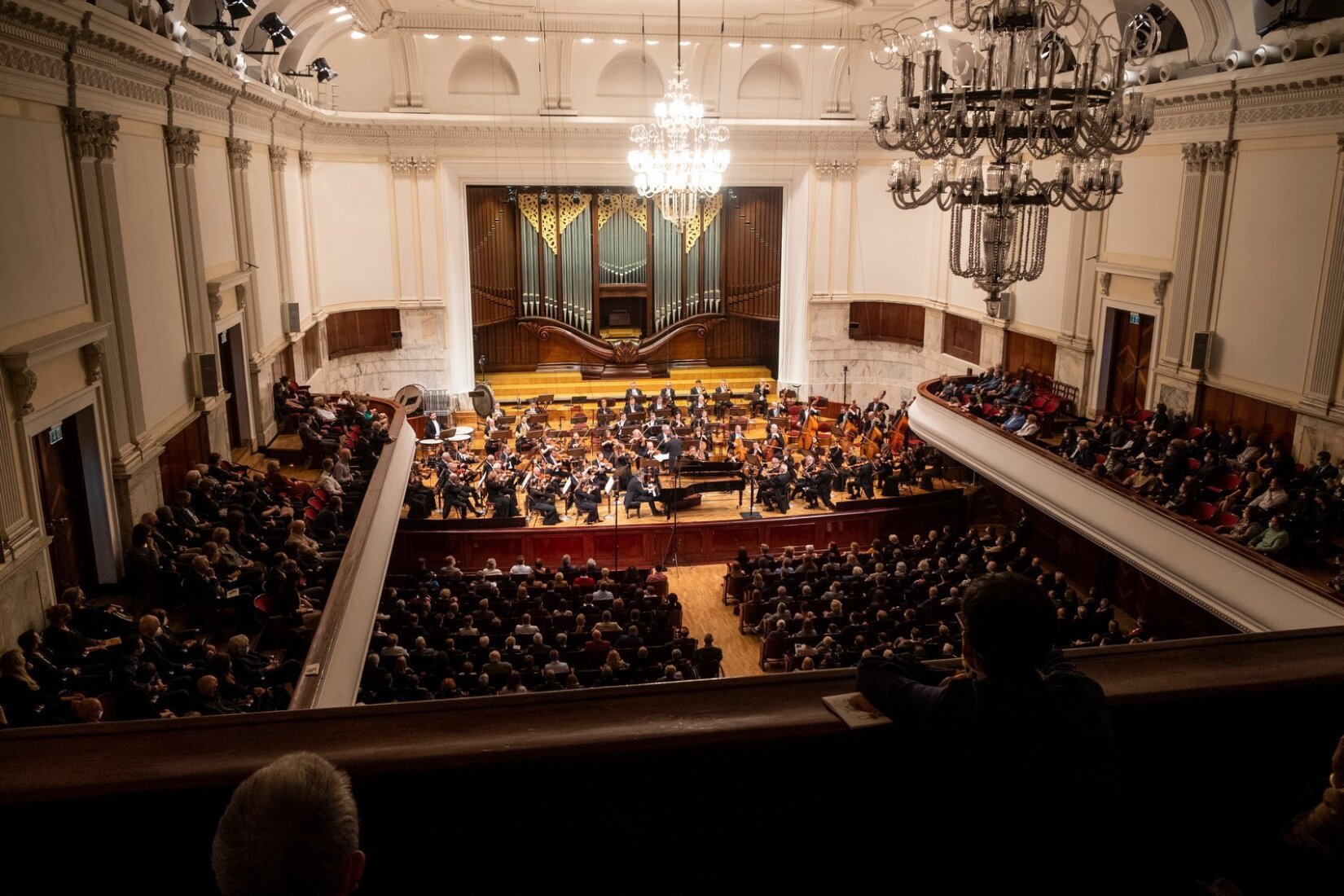 Fotografia przedstawia Salę Koncertową Filharmonii Narodowej w Warszawie. Sala jest ukazana z wysokości balkonu znajdującego się z tyłu sali. Na widowni oraz bocznych balkonach siedzi publiczność, na scenie orkiestra z instrumentami. W tle za orkiestrą znajdują się ozdobne organy. Na suficie dwa duże żyrandole.